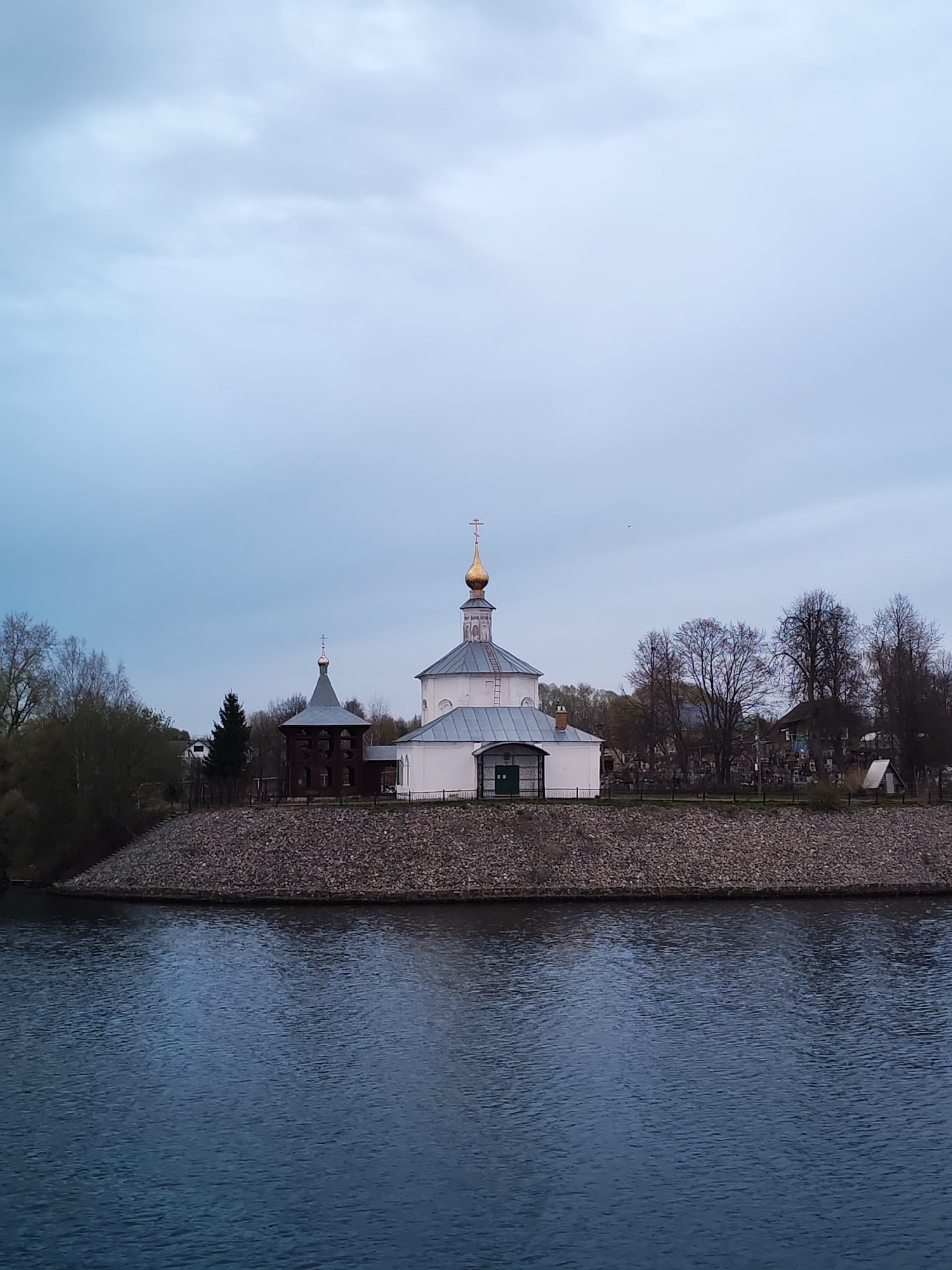 The image depicts a tranquil scene featuring the Church of St. Nicholas in Kopylovo, Russia. The church is the focal point, with its white walls, grey roof, and distinctive golden dome topped with a cross. Adjacent to the church is a smaller, darker wooden structure, possibly a bell tower, also with a spire and cross. The buildings sit atop a rocky embankment that slopes down to the water's edge. The water itself is dark and slightly rippled, reflecting the overcast sky above. Trees line the embankment, some still bare of leaves, suggesting it is either early spring or late autumn. The overall atmosphere is serene and somewhat muted due to the soft, diffused light from the cloudy sky.
