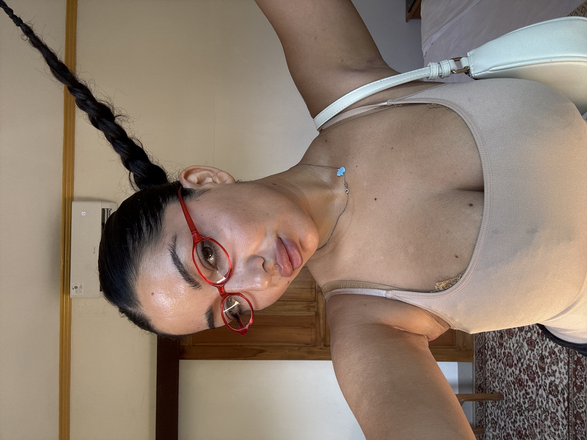 The image depicts a woman with dark hair styled in a long braid, wearing red-framed glasses and a beige tank top. She has a fair complexion and is wearing a delicate necklace with a light blue pendant. A white purse with a white strap is slung over her shoulder. The background includes a cream-colored wall and a wooden cabinet or dresser. Part of a patterned rug is visible in the lower right corner. The image appears to be taken indoors with artificial lighting. There is no discernible information that allows the location to be specifically identified as North Kuta, Indonesia.