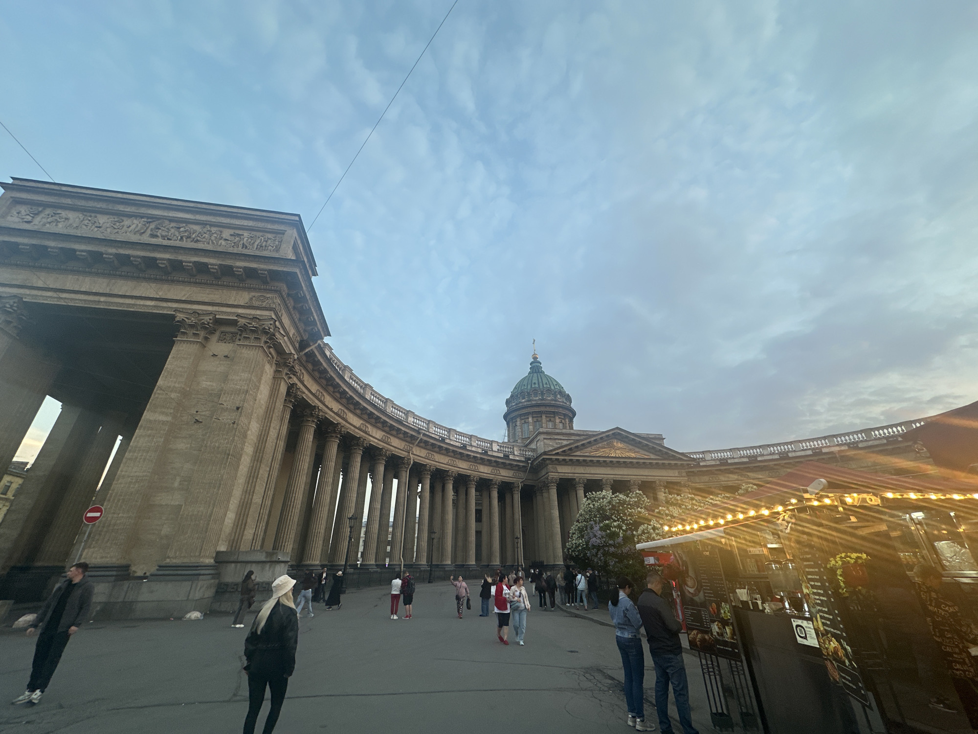 The image captures the Kazan Cathedral in Saint Petersburg, Russia. The cathedral's semi-circular colonnade dominates the scene, with the central dome visible in the background. Numerous people are walking around the square in front of the cathedral. There is a food stall with lights along the roof. The sky is overcast.