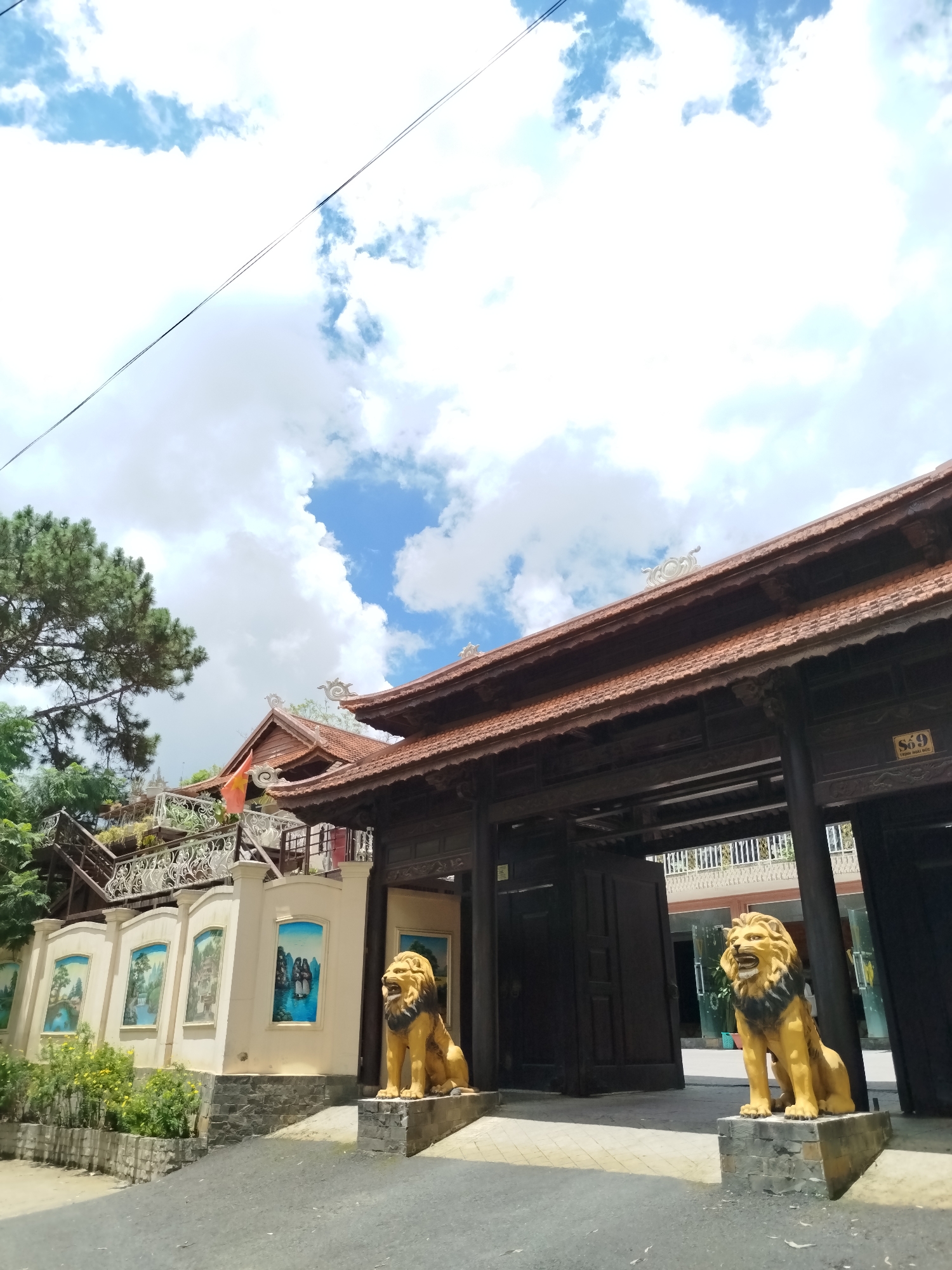 The image shows a facade of a traditional Vietnamese building in Xuân Trường - Đà Lạt, Vietnam. In the foreground, there are two golden lion statues flanking the entrance. The building has a dark wooden exterior with a tiled roof. To the left, there is a wall adorned with several paintings of landscapes and figures, and a staircase leading to an upper level with white railings and greenery. A tall pine tree is visible on the far left. The sky above is blue with scattered white clouds. The ground in front of the building is paved. The scene appears to be an entrance to a temple or a cultural site.