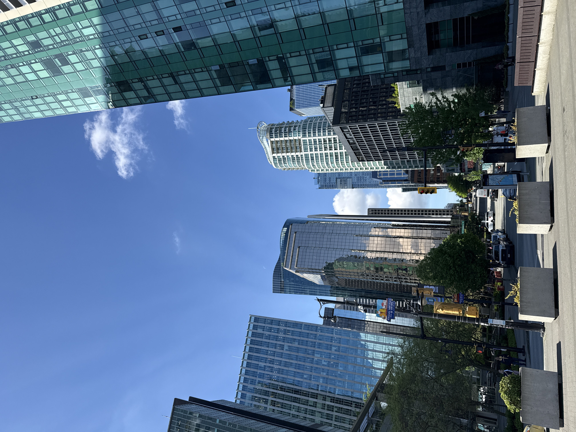 The image depicts a street scene in Vancouver, Canada, surrounded by modern skyscrapers. The perspective is from street level, looking upwards at the buildings and sky. The sky is blue with scattered clouds. Several tall buildings with glass facades reflect the sky and surrounding buildings. A street runs between the buildings, with sidewalks, trees, traffic lights, and planters. Several cars and pedestrians are visible on the street. A street sign that says "Howe" and "Melville" indicates the intersection of Howe and Melville Streets.