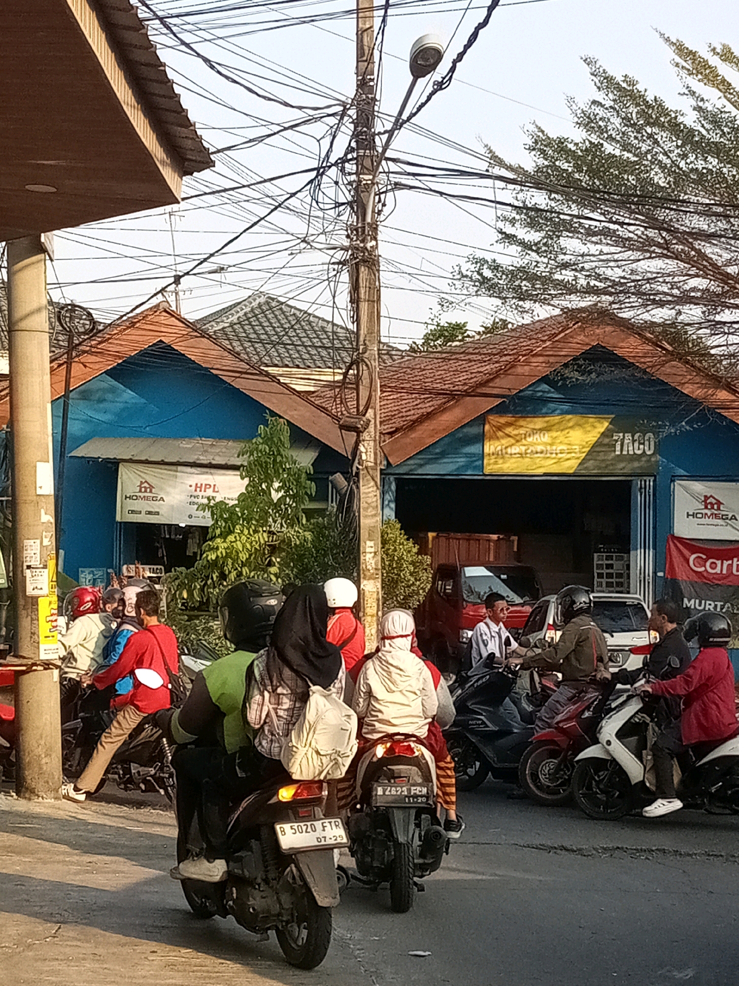 The image depicts a street scene in Tambun, Indonesia, on a sunny day. Multiple motorcycles are present, with riders and passengers wearing helmets. Some individuals are wearing casual clothing, while others are in uniforms. The background features buildings with blue facades and tiled roofs, adorned with signs for businesses like "HOMEGA HPL" and "TOKO MURTAHOFF 7ACO." A utility pole with numerous electrical wires is prominently positioned in the foreground, along with street lamps. The road surface is paved, and shadows suggest the sun is at a moderate angle. The overall atmosphere suggests a typical urban environment with active street life.