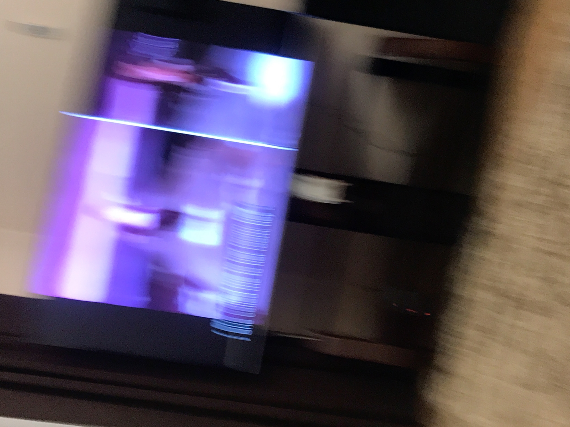 The image is blurry and difficult to decipher. It appears to show a television screen with a purple and white image displayed on it. The television is positioned on a dark brown cabinet or surface. To the right of the television, there is a blurry, light brown, textured surface that may be a wall or other object. Due to the blurriness, no people, specific objects, actions, or interactions can be identified. The image does not provide enough information to determine a location within the city of Yerevan, Armenia.