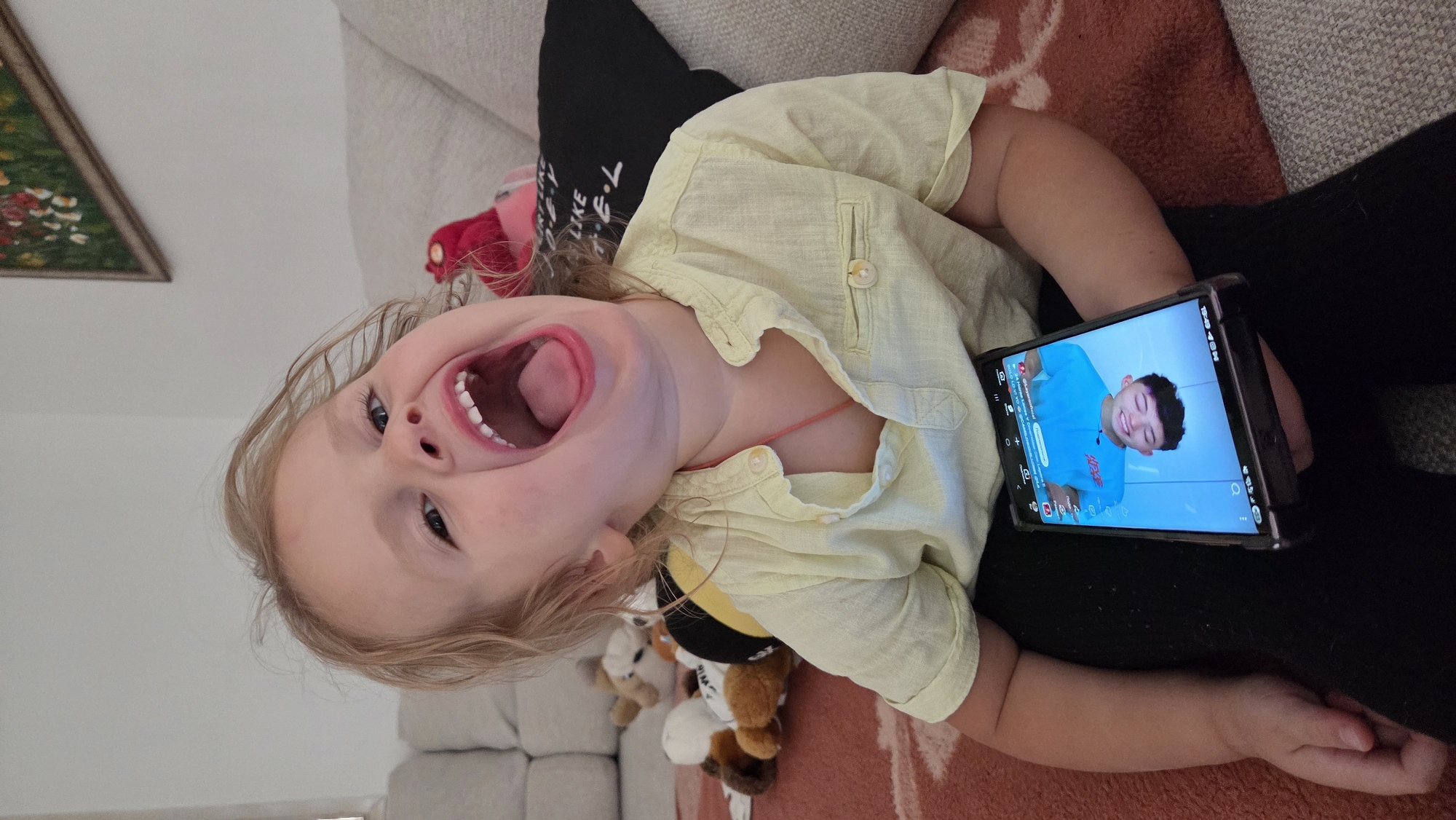 The image features a young child with blonde hair, wearing a light yellow short-sleeved shirt, lying down. The child's mouth is wide open in a laugh or shout, and their eyes are looking upwards. The child is holding a smartphone in their right hand, which displays a video of a young person with dark hair wearing a blue t-shirt. The background shows a couch and a textured blanket, suggesting an indoor setting. Several plush toys are visible near the child's torso, including one that appears to be a bee. A framed picture with greenery and flowers can be seen in the upper left corner. The lighting suggests it might be daytime. The overall emotion conveyed is one of joy and engagement. The image is oriented sideways.