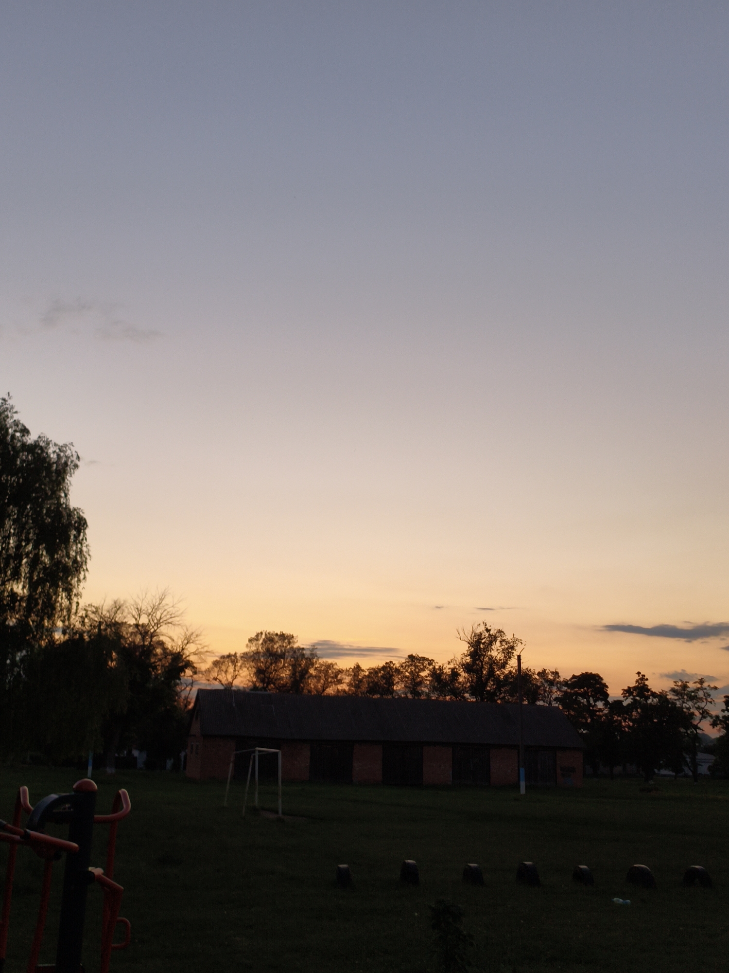 The image depicts an outdoor scene at dusk in Ustyvytsia, Ukraine. A brick building with a dark roof is centrally located, flanked by trees. In the foreground, a piece of outdoor exercise equipment with red and black components is visible on the left. A soccer goal is positioned on the grass in front of the building. The sky transitions from a light blue at the top to a soft orange hue near the horizon, indicating sunset. The overall atmosphere is calm and serene.