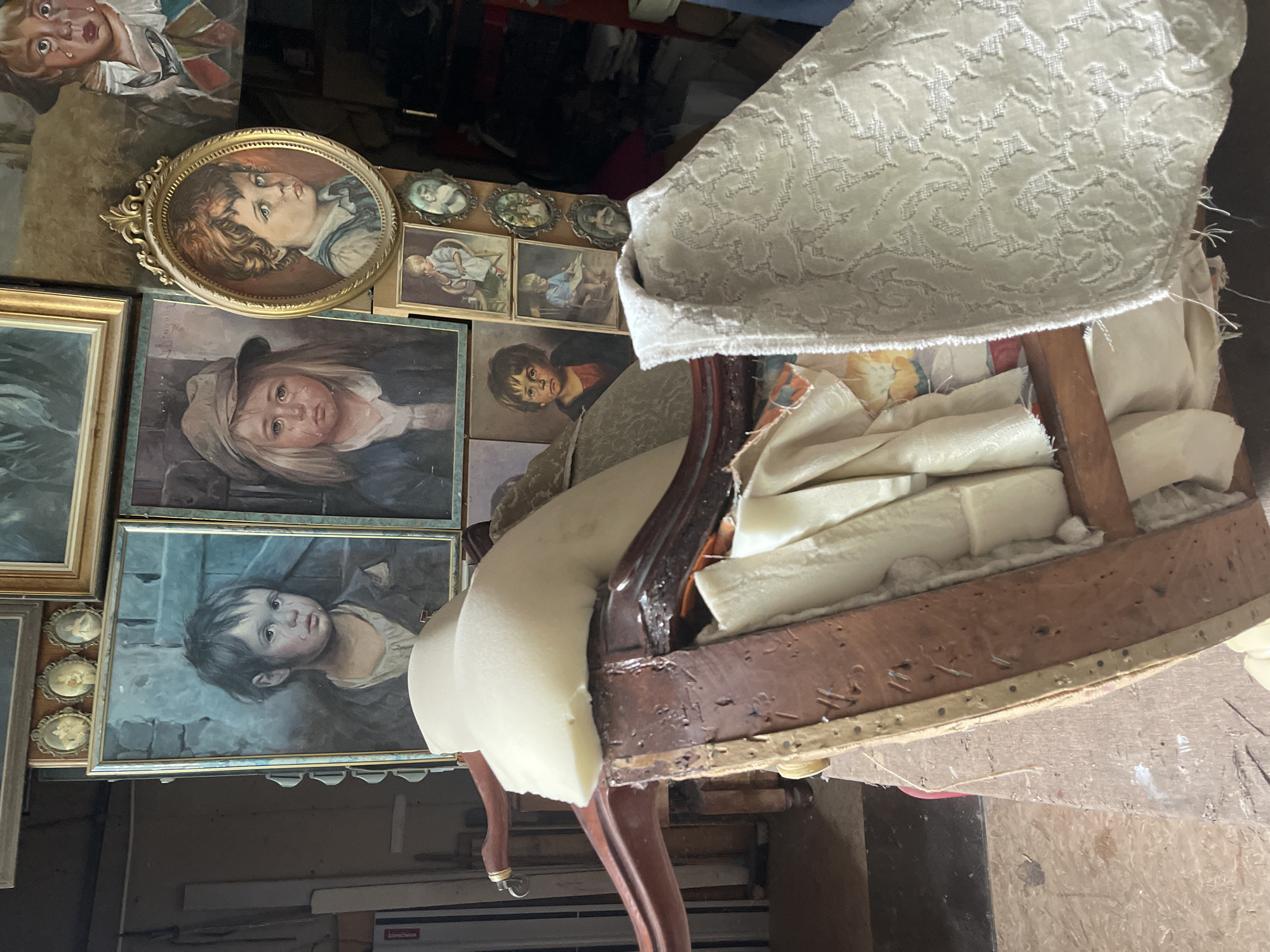 The image displays a cluttered indoor environment, possibly a workshop or storage area, filled with various framed paintings of children, a partially disassembled armchair, and stacks of fabric. The paintings are predominantly of sad-faced children, some in oval frames and others in rectangular frames. The armchair, made of dark wood, is in a state of disrepair, with exposed foam padding and wooden structural elements visible. Several pieces of patterned fabric are draped over the armchair and surrounding surfaces. The background is dimly lit and shows more indistinct objects, suggesting a space filled with stored items. The overall impression is one of an unfinished project or a collection of vintage items in a state of disarray. The scene is set in an unknown location within the city of Unknown.