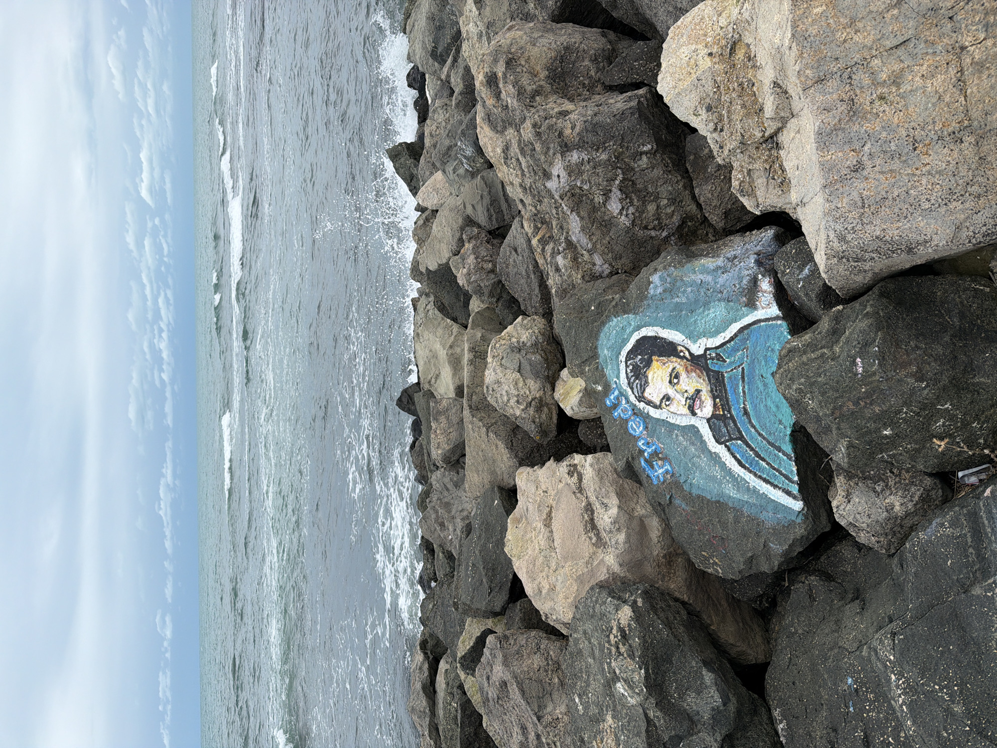 A weathered rock formation on a beach in Primorsko, Bulgaria, is adorned with a painted portrait of Freddie Mercury, the iconic lead singer of Queen. The portrait, rendered in vibrant blues and yellows, is located on one of the medium-sized rocks amidst the larger, grey boulders that make up the shore. The sea, with its choppy, grey waves, crashes against the rocks in the background, under a cloudy, overcast sky. The overall scene is one of a rugged, natural landscape marked by an unexpected artistic tribute.