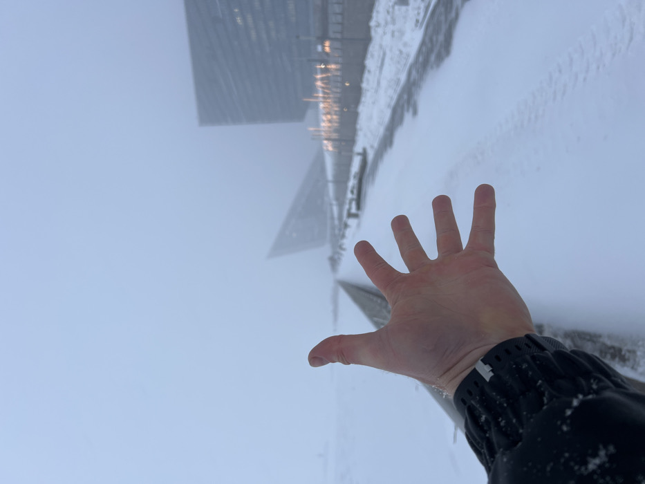 A first-person perspective shot depicts a winter scene at the Lakhta Center complex in Saint Petersburg, Russia. In the immediate foreground, a pale left human hand, fingers slightly splayed, extends towards the viewer from the lower center. The individual's dark, possibly black, jacket sleeve with textured fabric and a light-colored band on the cuff is visible at the bottom left. Snowflakes are visible on the jacket cuff.
The scene beyond the hand is covered in fresh snow. A wide, snow-covered path or embankment stretches from the midground into the distance on the right side of the frame. Faint imprints resembling tire tracks or footprints are discernible in the snow on this path. Parallel to the path on the left is a lower, dark barrier or railing, beyond which lies a vast, flat expanse of snow, likely a frozen body of water or another open area.
Through dense fog or heavy overcast conditions, two large, modern buildings are partially visible in the distant background. To the left, an angular, dark, triangular-shaped structure rises. To the right, a taller, more rectangular building with multiple illuminated windows is discernible. Rows of indistinct lights are visible along the embankment or adjacent structures in the mid-to-background, casting a faint glow. The sky is uniformly light grey, indicating low visibility and overcast weather. The overall environment is cold, snowy, and hazy.
