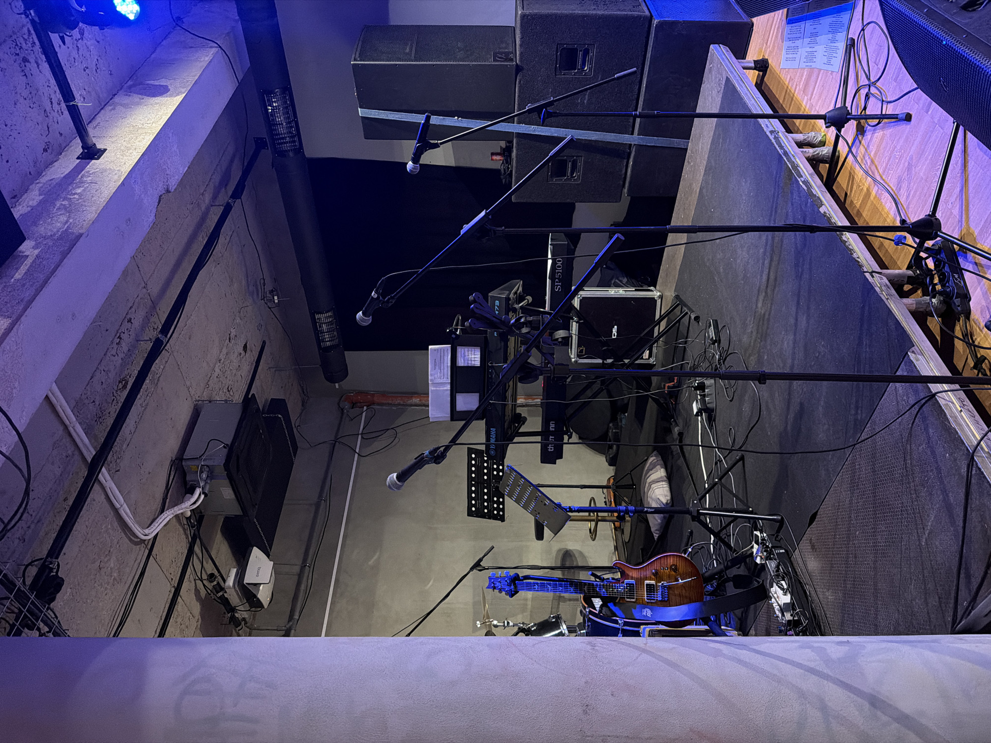 The image shows a stage or performance area in an urban location, featuring musical instruments, equipment, and infrastructure. Visible elements include microphones, speakers, a guitar, drum elements, and music stands. Various cables and electrical systems are routed along the walls and ceiling. The environment exhibits an industrial or urban aesthetic, with concrete surfaces and exposed utilities. Overall, the scene depicts a setup for a live music performance.