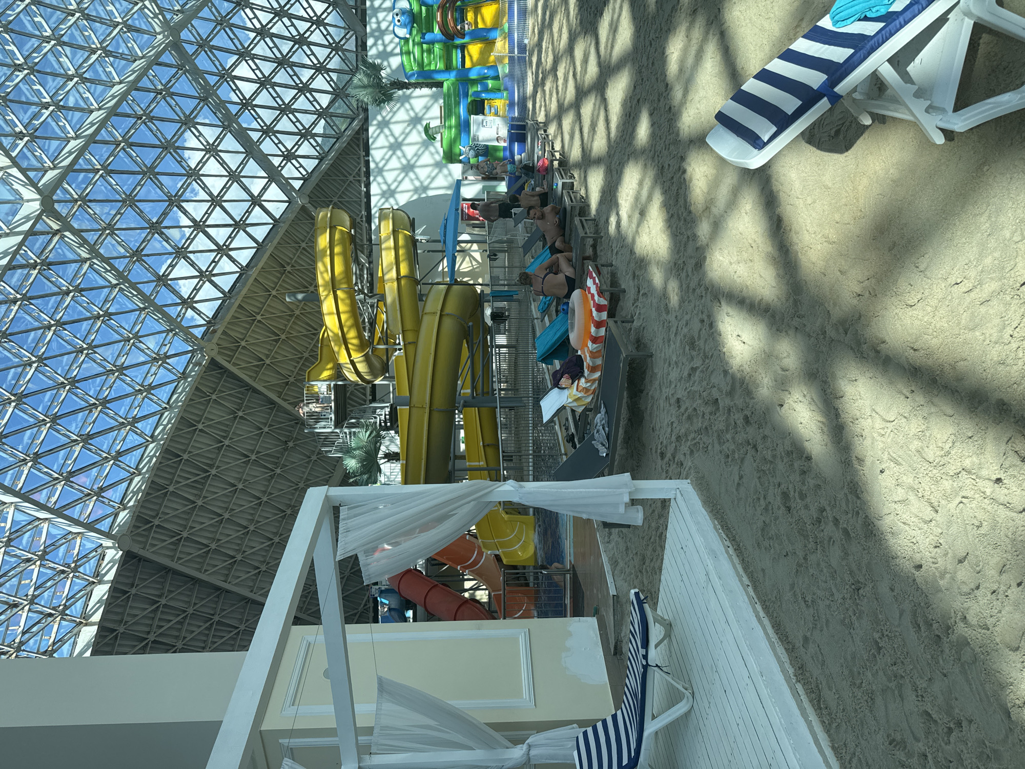 The image depicts an indoor water park with a sandy beach area. In the background, there are large yellow and red water slides, and a green inflatable water structure with an elephant motif. The ceiling is a geodesic dome with a metal framework and glass panels, allowing natural light to enter.
In the foreground, there are several lounge chairs on the sand. Some are occupied by people, including a woman in a bikini and a man lying down on a lounge chair. There are also towels and other personal items on the chairs. A white gazebo with sheer curtains is visible on the left. Another striped lounge chair is partially visible in the lower right corner of the image.
The location is identified as the city of Estosadok, Russia. The overall scene suggests a leisure and recreational environment.