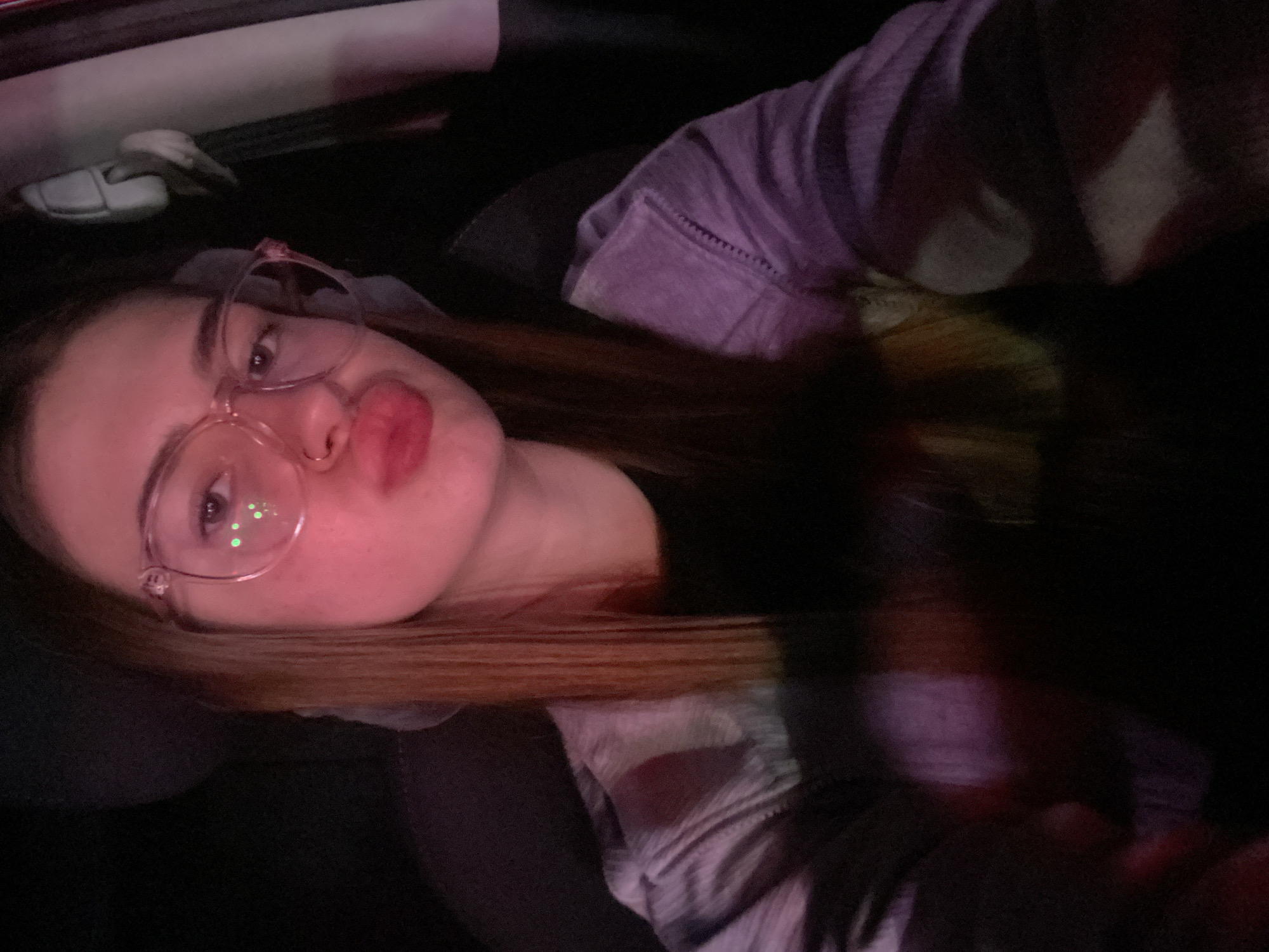 A young woman with blonde hair and glasses is sitting in a car. The woman is wearing a purple sweatshirt. The lighting is dim, with pinkish and purplish tones reflecting off her face and clothing. She is looking at the camera with her lips pursed. The background is dark and indistinct, suggesting it is nighttime or the interior of the car is dimly lit. The location is not identifiable within the image itself, therefore the mention of Дьяково, Russia is not supported by visual evidence.