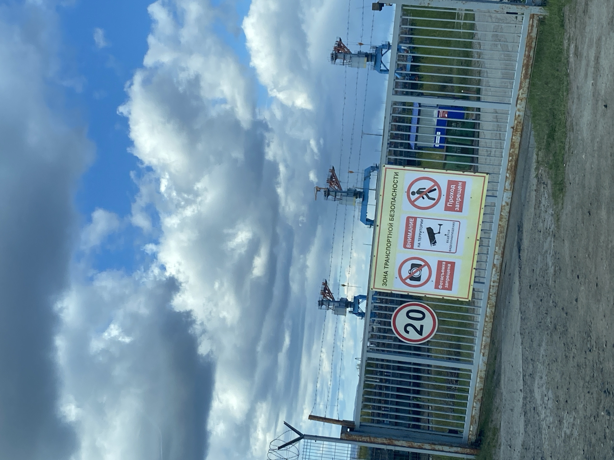 A slightly rotated medium shot shows a fenced-off area under a cloudy sky, identified by a sign in Russian that says, "TRANSPORT SAFETY ZONE". The fence is tall, made of metal, and topped with barbed wire. A sign attached to the fence warns, "No Entry" and "No Photography Allowed". A speed limit sign with "20" inside a red circle is also present. Above the fence, three industrial cranes are visible against the sky. The surrounding area appears to be industrial with some green vegetation. The location is likely in or near the city of Chaikovsky, Russia, based on the language of the sign.