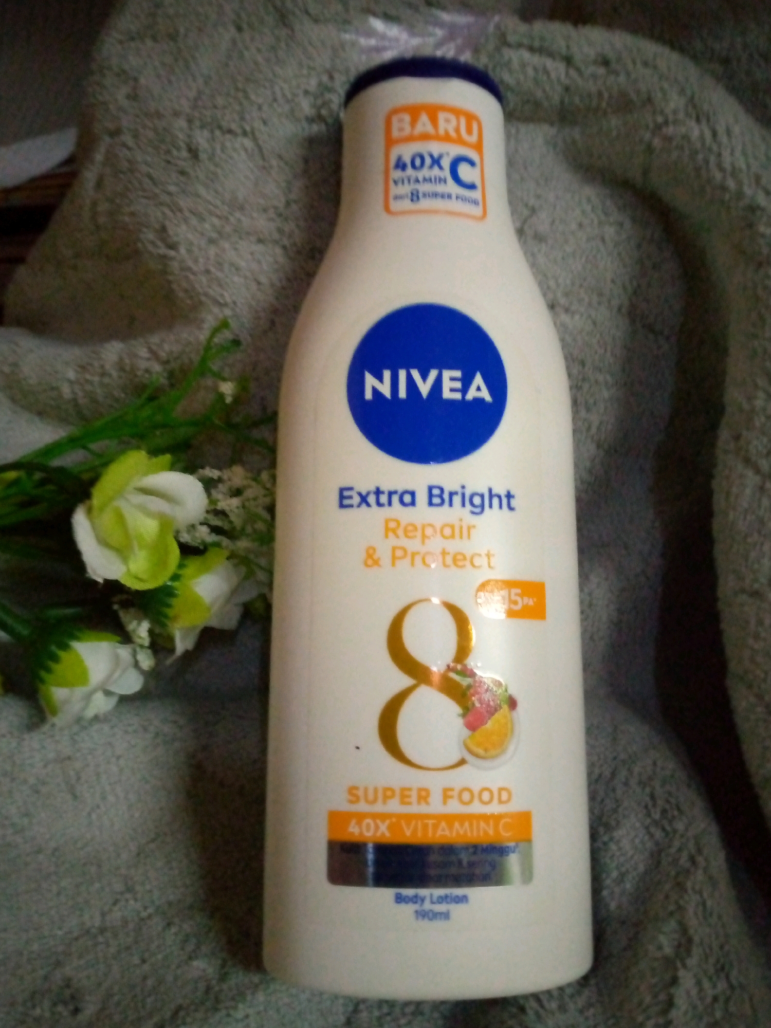 A white Nivea body lotion bottle is placed on a textured gray fabric surface, with a few artificial white and green flowers to its left. The bottle features prominent branding and text, including "BARU 40X VITAMIN C & 8 SUPER FOOD" in orange and blue, "NIVEA" in a blue circle, and "Extra Bright Repair & Protect" in blue and orange. A large golden number "8" with fruit illustrations is also visible on the bottle, along with "SUPER FOOD 40X VITAMIN C" and "Body Lotion 190ml." The lighting is dim, casting a shadow on the right side of the bottle. The scene does not provide any contextual information or visual cues to suggest its location within the city of Sumberwono, Indonesia.