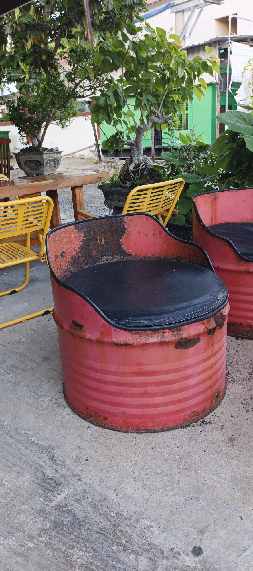 The image captures an outdoor seating area, likely in Phu Thuy Ward, Vietnam. The foreground is dominated by two chairs made from repurposed red oil drums with black cushioned seats. These chairs exhibit signs of wear and rust, particularly on their exteriors. To the left of the chairs, a yellow metal chair with a slatted design is visible. Behind the seating arrangement, a wooden table stands, accompanied by more yellow chairs.
The background reveals lush greenery from trees and plants, some of which appear to be bonsai. Buildings with green and white facades are present, suggesting an urban or semi-urban environment. The ground is paved with concrete, showing some wear and dirt. The overall scene depicts a rustic and casual outdoor space, possibly a cafe or a residential patio, with a focus on recycled materials for furniture.
