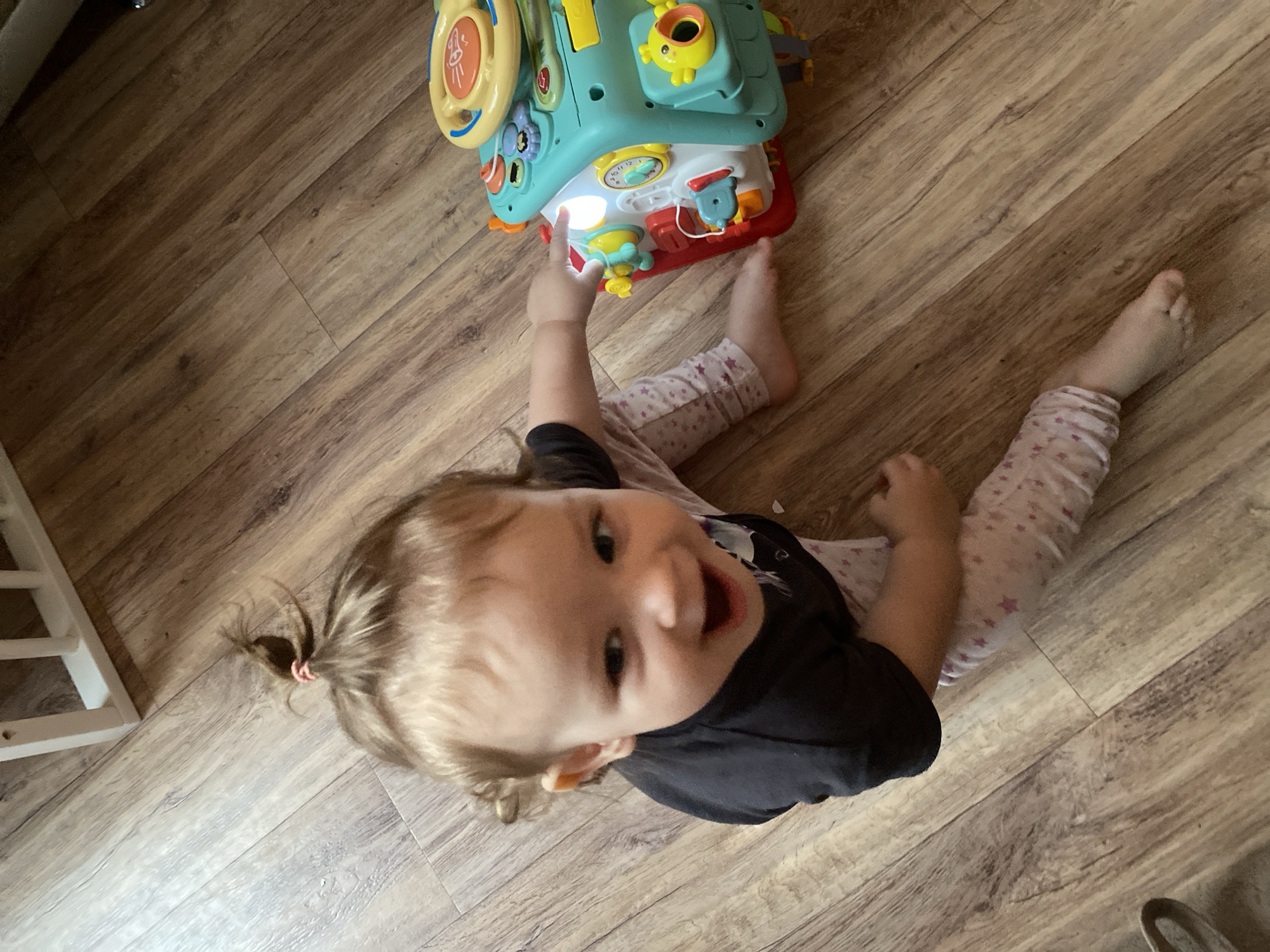 A high-angle shot captures a young child sitting on a wooden floor, looking directly at the camera with a wide, joyful smile and an open mouth. The child has light-colored hair, styled in a small ponytail secured with a pink elastic band. They are dressed in a dark t-shirt and light-colored pants featuring a subtle star pattern.
In front of the child is a multi-functional, brightly colored activity toy, primarily teal and light blue, with accents of yellow, red, and orange. One section of the toy emits a bright white light, and the child's right hand is extended towards it, suggesting active engagement with the toy. The child's bare feet are visible, splayed out on the floor behind the toy.
The setting is an indoor space with a light to medium brown wooden floor, featuring distinct grain patterns. A small portion of a white structure, possibly a crib or playpen, is visible on the far left. The overall impression is one of playful interaction and happiness in a well-lit domestic environment. No discernible text is visible in the image.