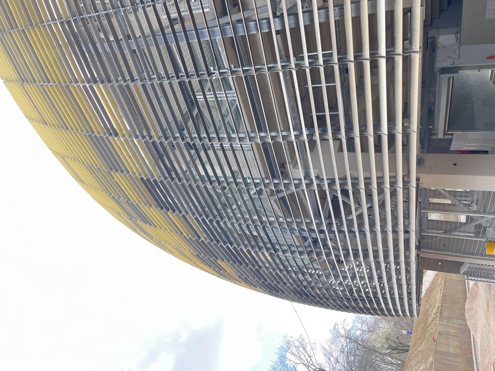 The image depicts a low-angle view of an exterior facade of a building under construction. The facade is composed of a dense grid of parallel, horizontally oriented metal slats, some of which are painted yellow. The slats provide shading. The architectural style appears modern and functional. The environment consists of a construction site, with bare earth and scaffolding visible along the lower right. Some deciduous trees with bare branches are present, suggesting winter or early spring. Based on architectural similarity and location data, the scene is potentially located in Sofia, Bulgaria. The building is under construction. No people are visible in the frame.