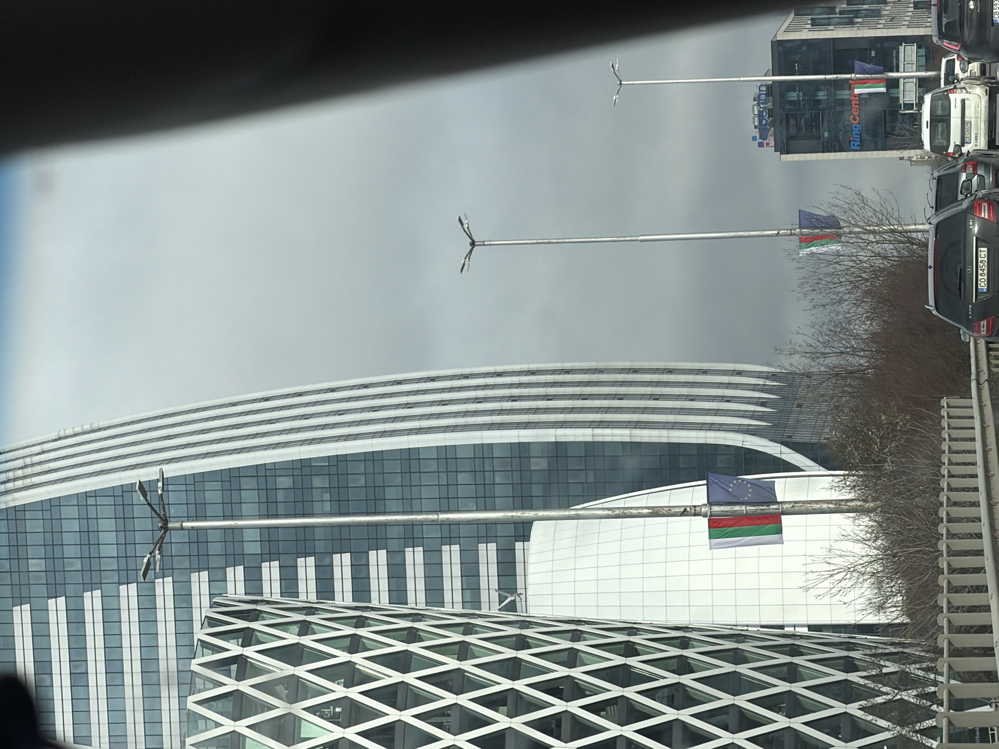 The image depicts a street scene in Sofia, Bulgaria. Prominent architectural features include modern buildings with curved and geometric designs, one with a diamond-patterned facade. Flagpoles displaying the flags of the European Union and Bulgaria are visible. In the background, taller buildings are also noticeable. In the foreground, there is traffic and parts of a vehicle, indicating the picture was taken from inside a car. The sky appears overcast.