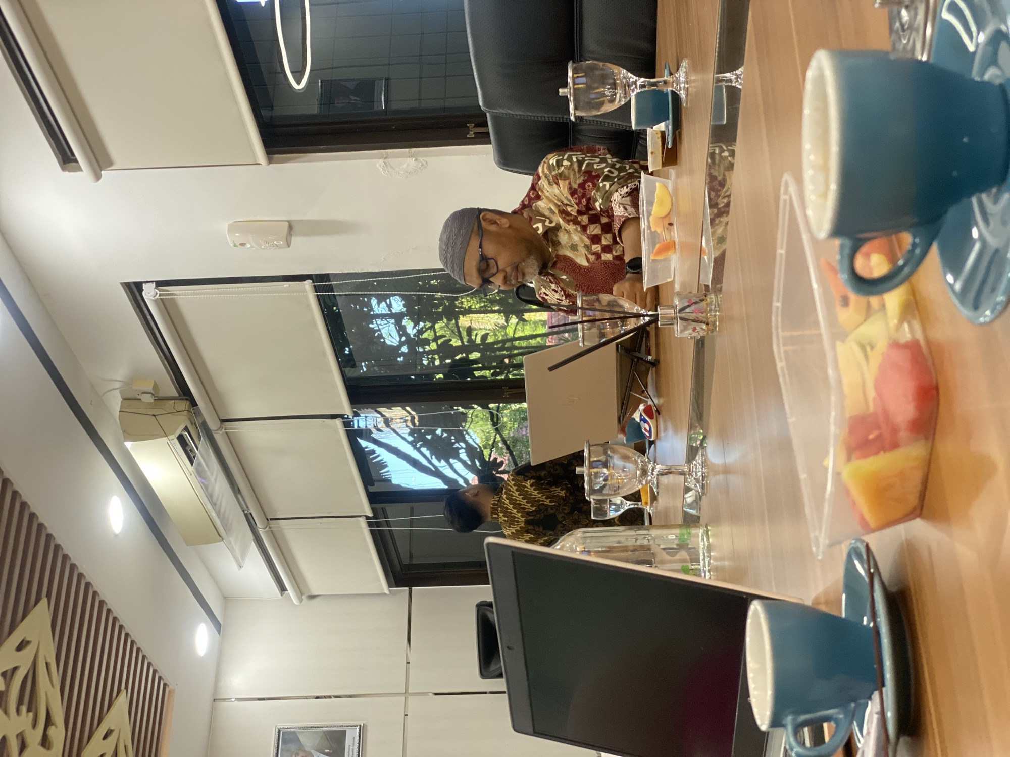 The image shows an indoor scene, likely a meeting room in Banjarbaru, Indonesia. In the foreground, a wooden table holds two blue coffee cups with saucers, a container of cut fruit, and a laptop. Further back, two men are seated at the table, facing a window. One man, wearing glasses and a grey skullcap, is in the right foreground, looking down. He is wearing a patterned shirt. To his left, and slightly further back, another man in a patterned shirt is visible from the back, looking out the window. The room features an air conditioning unit mounted high on the wall, and roller blinds covering the windows. The ceiling has a decorative wooden element. The overall ambiance suggests a professional or business setting.