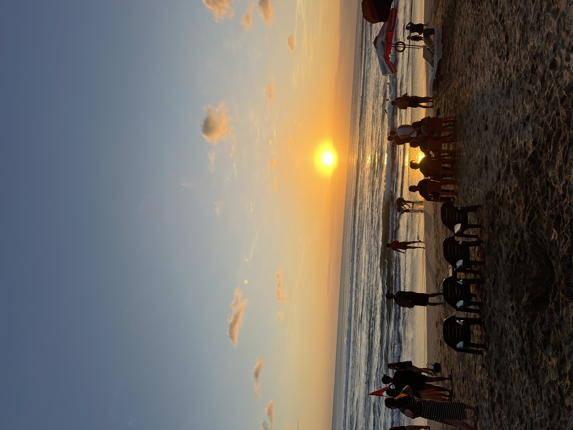 At sunset on Kuta Beach in Bali, Indonesia, a group of people are gathered on the sandy shore. The sun, a bright orange orb, is low on the horizon, casting a warm glow across the sky and the ocean waves. Several individuals are standing near the water's edge, some appear to be wading, while others are observing the sunset. A few groups of people are clustered on the beach, with some seated in plastic chairs. One person in the foreground is wearing a striped dress, and another is holding a red flag. A beach umbrella with a red and white pattern is visible on the right side of the image. The sand is textured with footprints and the gentle slope of the beach leads to the ocean, where waves are rolling in. The overall scene depicts a serene evening at a popular tourist destination.