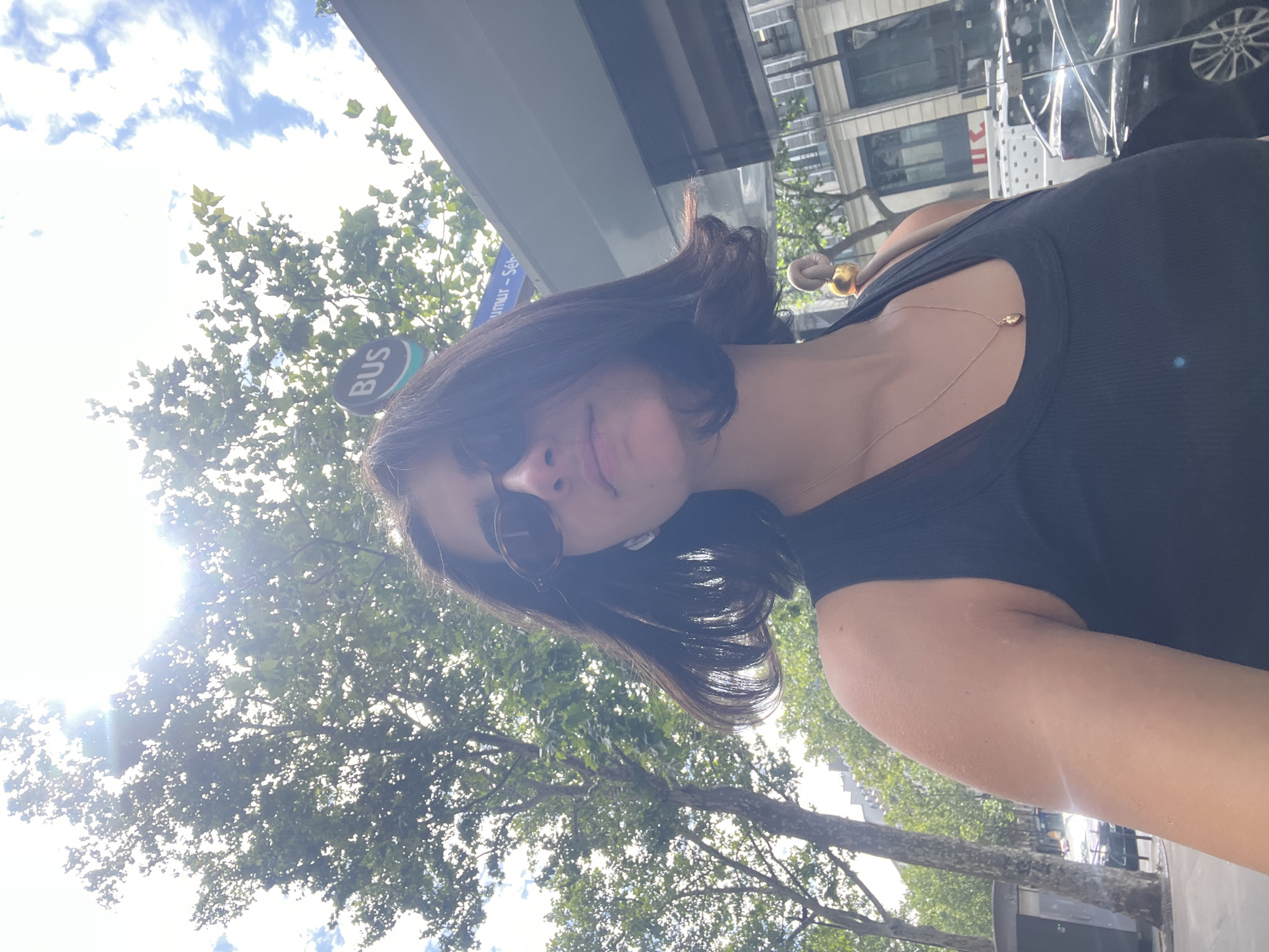 An individual with dark, shoulder-length hair and sunglasses is seen from the chest up, looking slightly upwards and to the right. They are wearing a black tank top and a delicate gold necklace. Behind them, a street scene unfolds in Paris, France, with trees, a bus stop sign that reads "BUS," and buildings in the background. The sun is shining brightly, casting lens flares and illuminating the sky with scattered clouds. Cars are visible on the street.