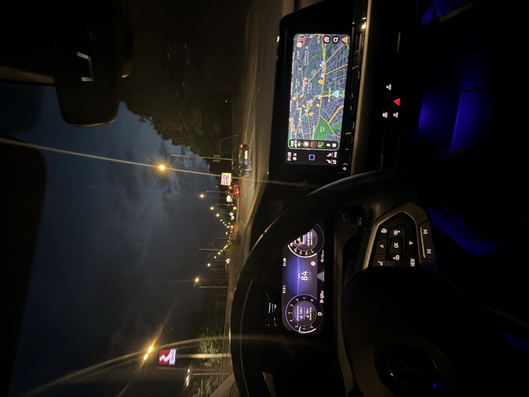 The image provides a first-person perspective from inside a car driving at night. The scene is illuminated by the car's interior lights and numerous streetlights outside.
Within the car's interior, a digital instrument cluster is visible through the steering wheel. It displays a speed of 84 (likely km/h), the gear selector in 'D', an estimated range of 220km, and an external temperature of 21.5°C. The oil temperature is shown as 100°C and coolant temperature as 90°C. The time on this cluster is 21:38. Additionally, text "Само собо" and "Jakone & YAGMI" are present, suggesting music information. To the right, a large infotainment screen shows a detailed navigation map with Cyrillic text, confirming the location as Sofia, Bulgaria. This screen also displays a time of 21:30 and "9/30," along with the word "SYNG."
Part of the steering wheel's right-hand controls are visible, including buttons for "OK," "VIEW," directional arrows, and volume. On the center console, the hazard light button (red triangle) is illuminated, along with other controls. Blue ambient lighting casts a glow on the car's interior, particularly in the lower right area.
Through the windshield, a multi-lane road is visible, brightly lit by a continuous line of streetlights stretching into the distance. The taillights of another vehicle are seen further down the road. On the left side, a prominent red sign with a white "M" symbol, commonly indicating a metro station, is visible, alongside another sign with Cyrillic text "ОНЛЕ." The sky is dark, indicating it is nighttime.