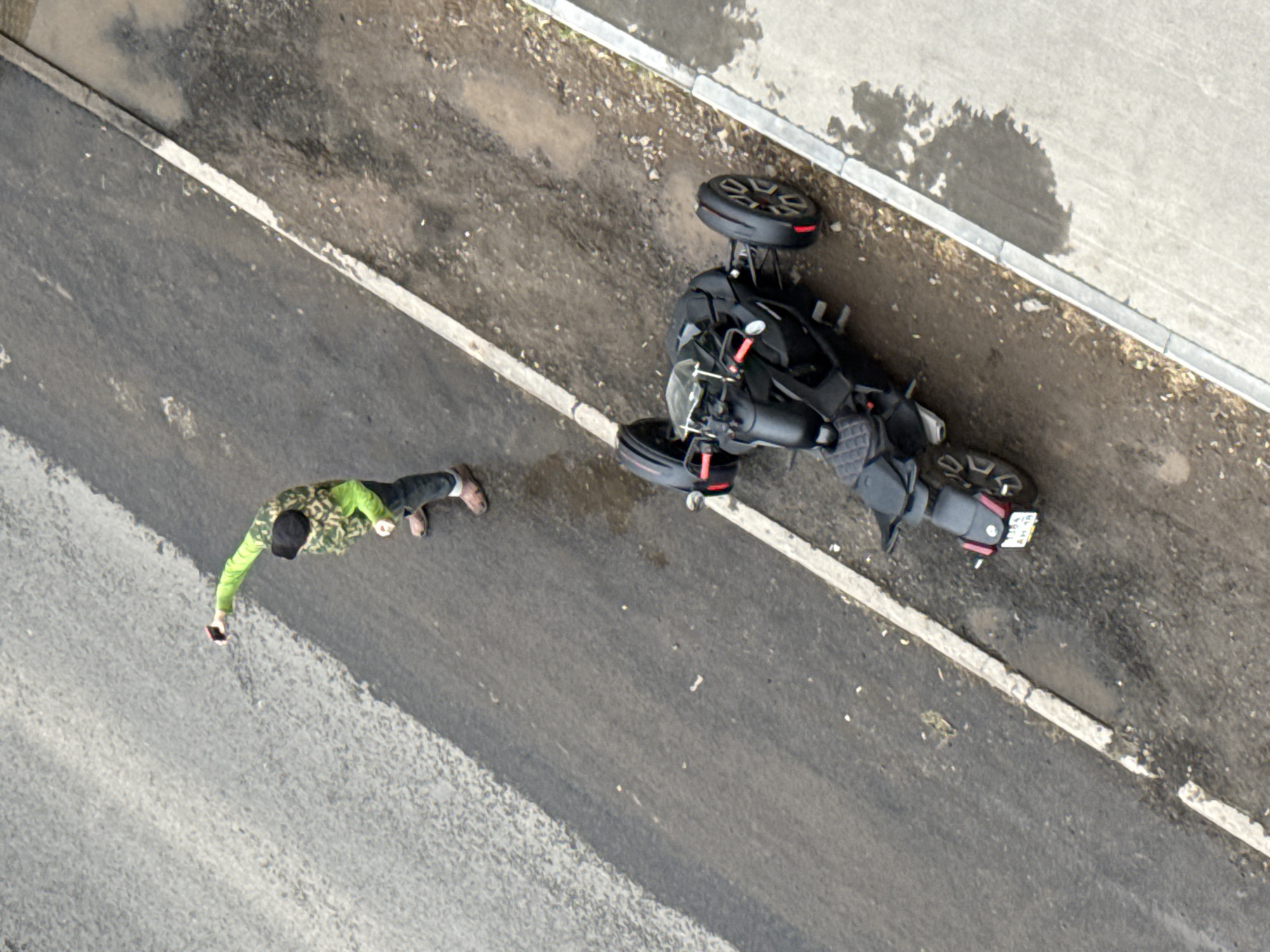 An aerial view shows a man with a dark hat, wearing a green and brown camouflage jacket and tan shoes, holding a cell phone while standing on the asphalt of a city street in Votkinsk, Russia. A three-wheeled motorcycle is lying on its side on the shoulder of the road near a concrete curb separating the road from a sidewalk, with leaves and debris visible near the curb. The street is wet in places. The license plate of the motorcycle is barely visible.