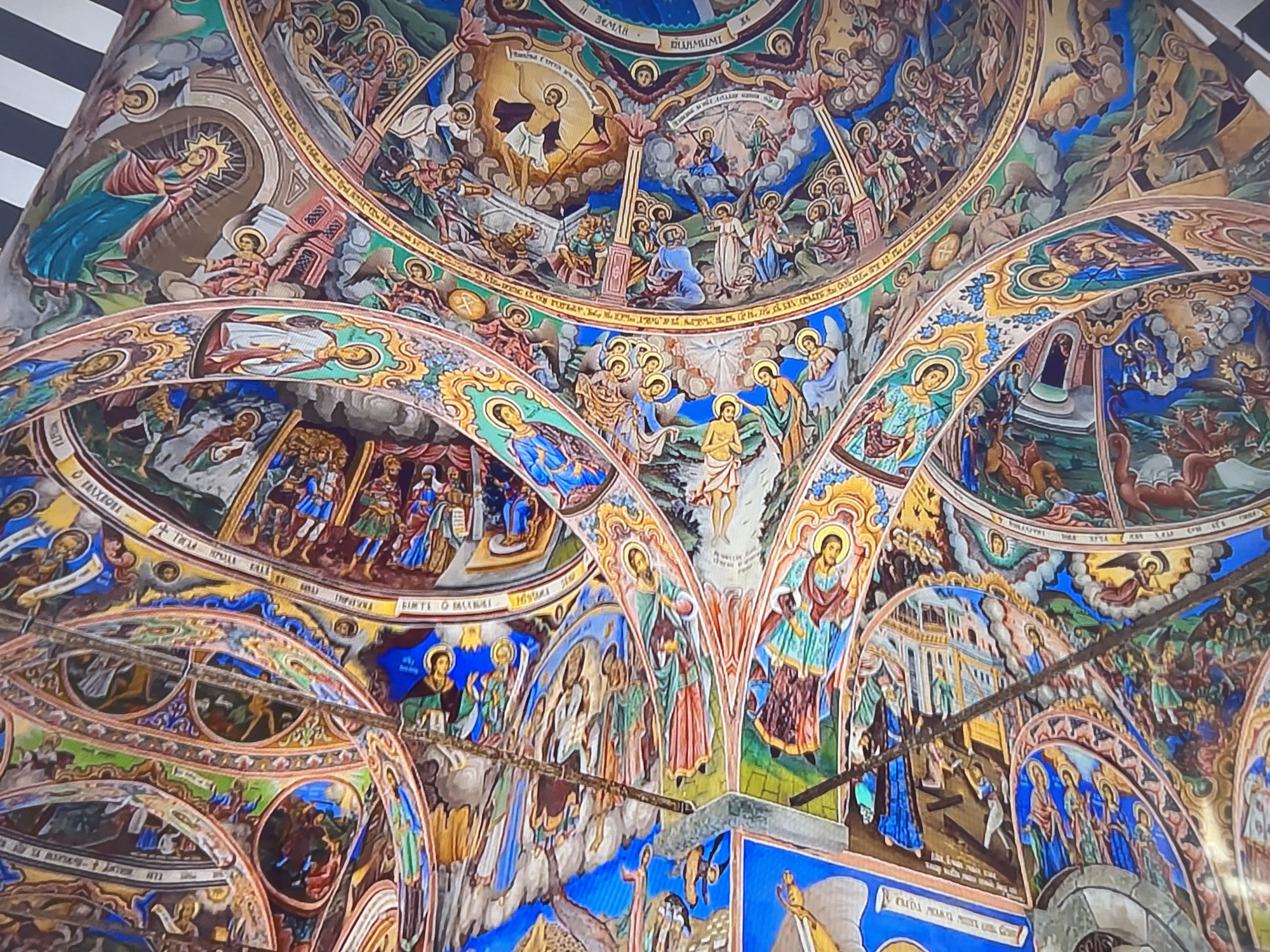 The image shows the interior of a church, possibly the Boyana Church near Sofia, Bulgaria, renowned for its medieval frescoes. The scene depicts various religious figures and biblical events painted on the walls and ceiling in a vibrant and detailed style. The frescoes feature numerous human figures, likely saints, angels, and scenes from the life of Christ, rendered in the Byzantine style. The environment consists of arched ceilings and walls covered in colorful and ornate paintings.