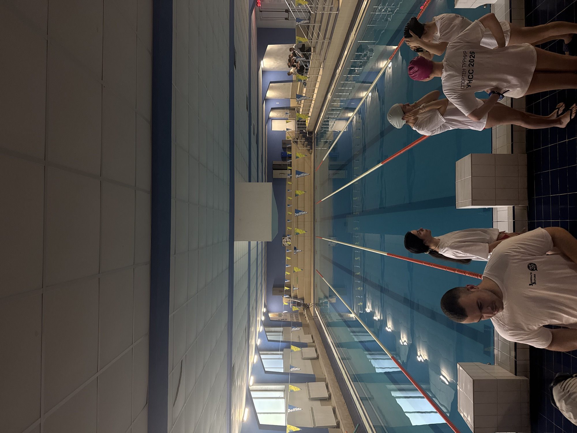 The image depicts an indoor swimming pool facility, likely during a sports event or practice session. The perspective is taken from above, looking down into the multiple lanes of the pool.
Several individuals, mostly young adults, are visible. In the foreground, a man and a woman stand by the starting blocks at the edge of the pool. They are wearing white t-shirts over their swimsuits. Further down the pool, three other women are also positioned at separate starting blocks, similarly dressed in white t-shirts, swimming caps (one pink, others light-colored), and goggles. In the blurred background, on the opposite side of the pool, a group of people can be seen, possibly spectators or other participants awaiting their turn.
The pool features clearly marked lanes separated by ropes, with starting blocks at one end. Yellow triangular flags, possibly advertising banners, are strung across the pool, bearing the text "Delfina", "fine", and "Ifine". The walls and ceiling of the facility are bright, featuring light-colored panels with blue accents. A digital clock in the background shows "10:1", potentially indicating 10:01 AM or PM.
The visible text on the white t-shirts confirms the event's nature: "СПОРТЕН ТУРНИР УНСС 2026" (SPORTS TOURNAMENT UNSS 2026), suggesting a university sports tournament, likely hosted by the University of National and World Economy (UNSS) given the Sofia, Bulgaria context. One man's shirt also bears the text "35 ЗАВИНАГИ ЗДРАВИ" (35 FOREVER HEALTHY), alongside a small logo. The scene conveys an atmosphere of anticipation or preparation for a swimming competition.