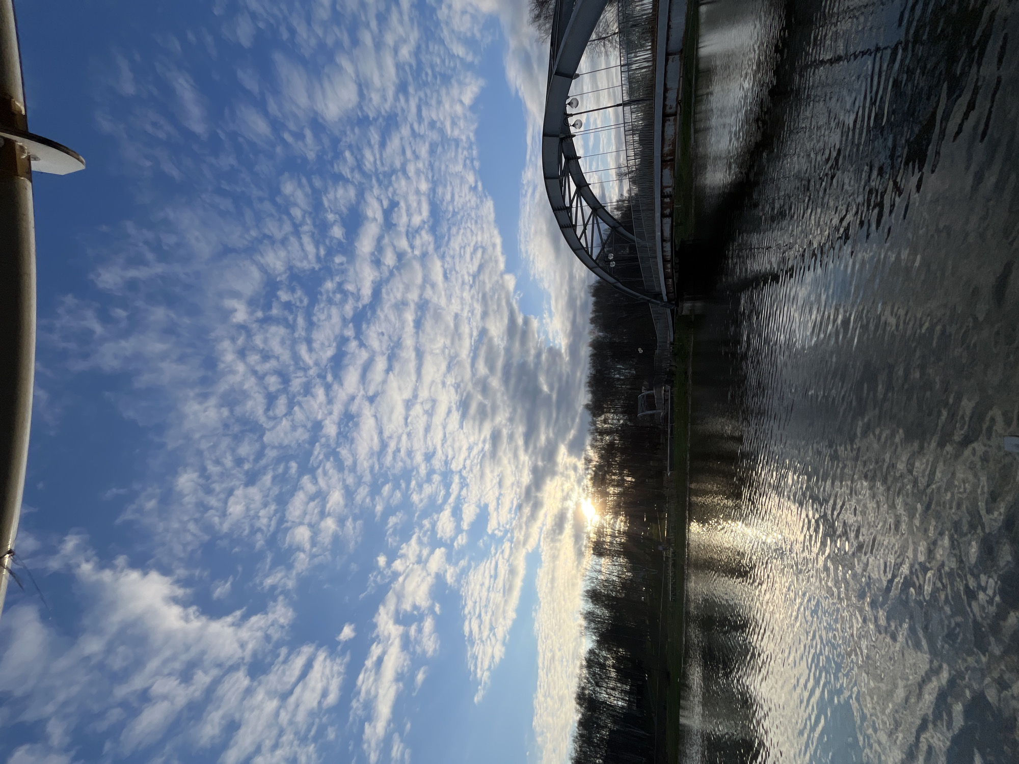 The image depicts a scene in Klin, Russia, featuring a body of water (likely a canal or river) reflecting the sky. A steel arched bridge spans the waterway, its structure partially visible on the right side of the frame. The sky is filled with altocumulus clouds. Trees line the bank of the water in the center of the image where sunlight is breaking through the branches. A lamp post appears in the lower left corner. The image seems to be rotated.