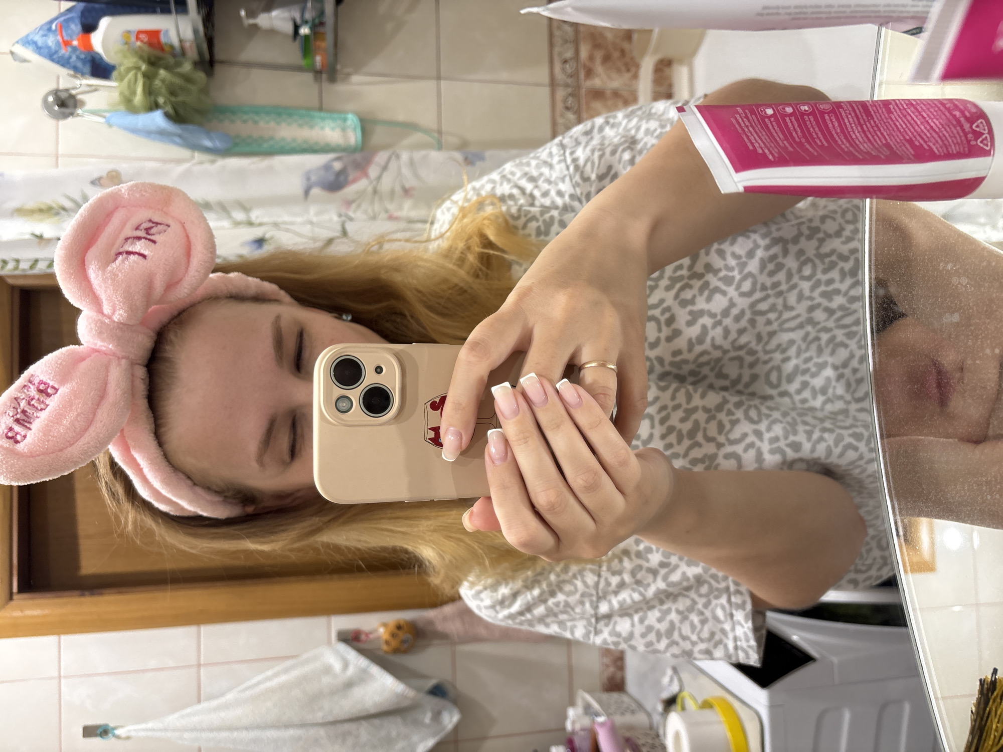 A woman in a bathroom mirror selfie. She wears a pink bunny-ear headband with "BOMB" embroidered on one ear. Her blonde hair is loose. She holds a beige iPhone with a red sticker of a heart with a stem and leaf. She has French-manicured nails and a gold ring on her finger. She wears a gray and white leopard-print shirt. The bathroom has white tiles and a wooden-framed mirror. Shelves above the mirror hold toiletries. A white towel hangs in the lower left. Below the counter are other items. It is not possible to determine the location to be Rusanovo, Russia from this image alone.