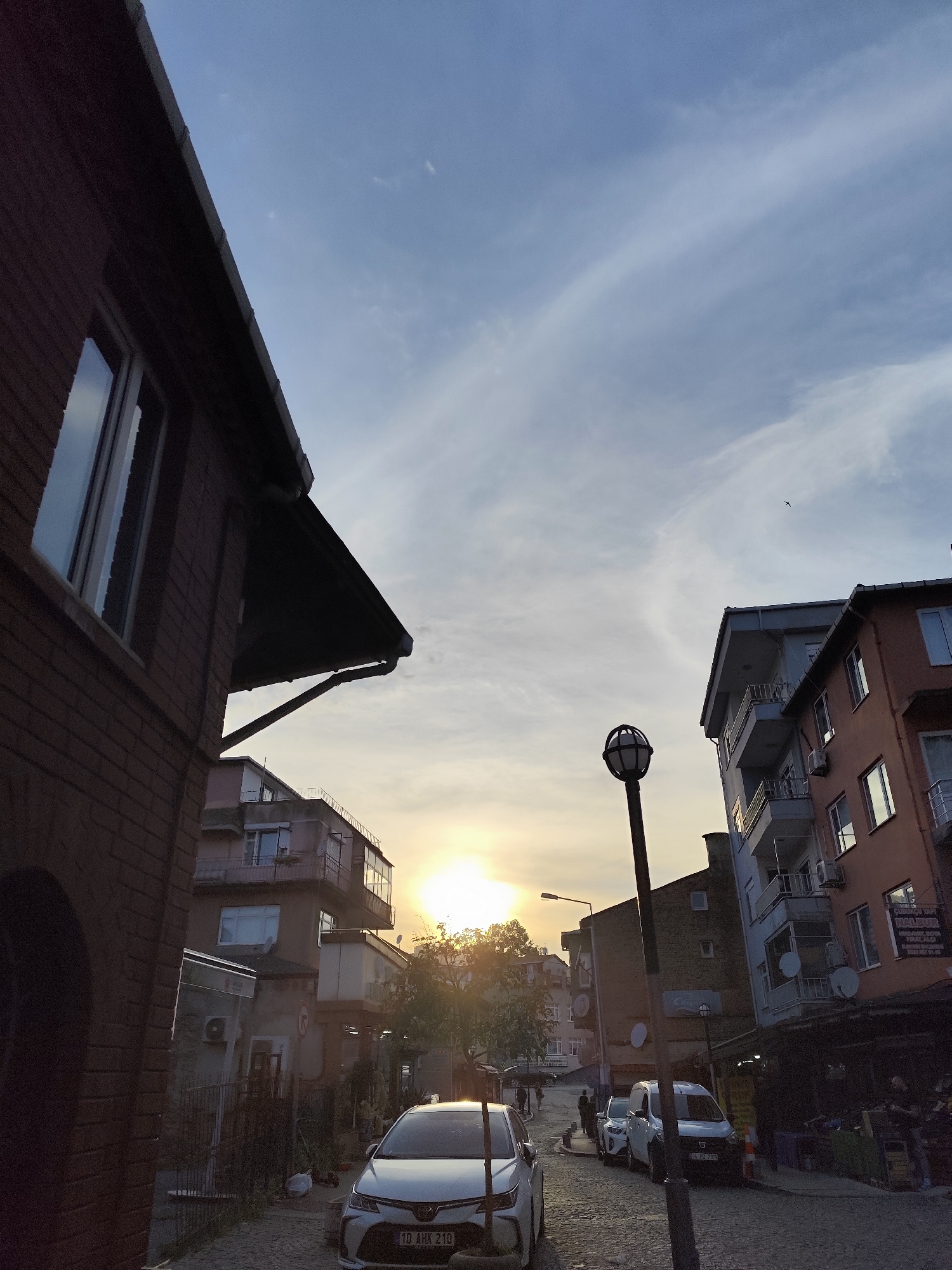 This image captures a vibrant street scene in Sarıyer, Turkey, at sunset or during the golden hour. The low sun is prominently visible on the horizon, casting a warm, golden glow across the cobblestone street and buildings, creating strong backlighting and a peaceful atmosphere. The sky above is partly cloudy, with wispy white clouds contrasting against the blue.
Multi-story buildings line both sides of the street. On the left, a brick building with a large window and a dark roof overhang is in the foreground. On the right, apartment buildings with balconies and air conditioning units are visible, some appearing older than others. A distinctive black street lamp with a spherical, caged light fixture stands prominently on the right.
Several vehicles are present, including a white Toyota Corolla sedan parked in the foreground with the license plate "1D AHX 210". Further down the street, a white van with the license plate "34 PD 799" and other cars are parked or moving. A few figures of people can be seen walking in the distance, silhouetted by the sun. Vegetation, likely trees and bushes, is also visible, particularly around the sun's position.
Visible text includes the license plates mentioned. On a building on the right, signage partially reads "CUMHURIYET YAPI MALZEMELERI" with phone numbers, suggesting a building materials store. A faint Coca-Cola logo is also visible on a wall further down the street.