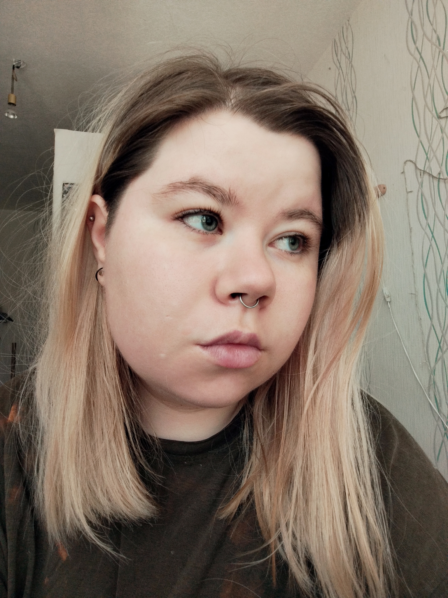 A close-up, eye-level, medium shot photograph depicts a young, fair-skinned individual with light brown hair transitioning to blonde ends. The person is looking off to the right of the frame. They have a septum piercing and two piercings on their left ear; a helix and a lobe piercing with a small hoop earring. The person is wearing a dark, possibly tie-dyed, t-shirt. The background features a light-colored wall with a subtle, curving pattern, and a glimpse of a white doorframe and a ceiling light fixture at the upper left.