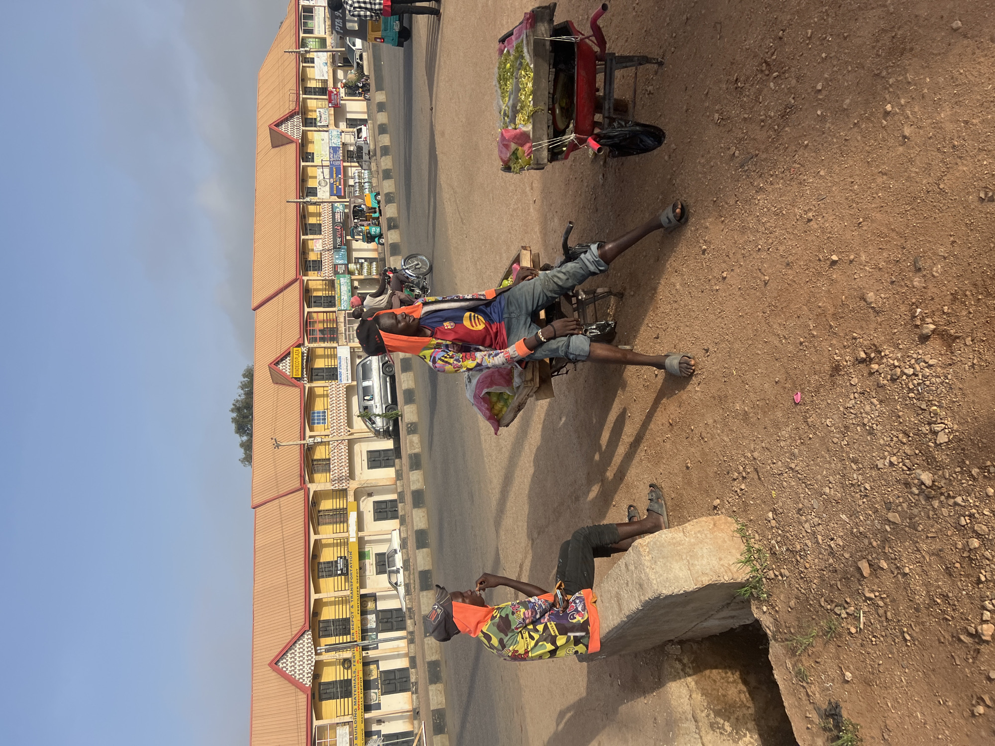 Two men rest on the roadside in Gura Top, Nigeria. One man reclines on a handcart loaded with goods, wearing a soccer jersey, shorts, and sandals. The other sits on a concrete ledge, dressed in a colorful patterned top and shorts. Buildings line the opposite side of the road. The ground is dirt and gravel.