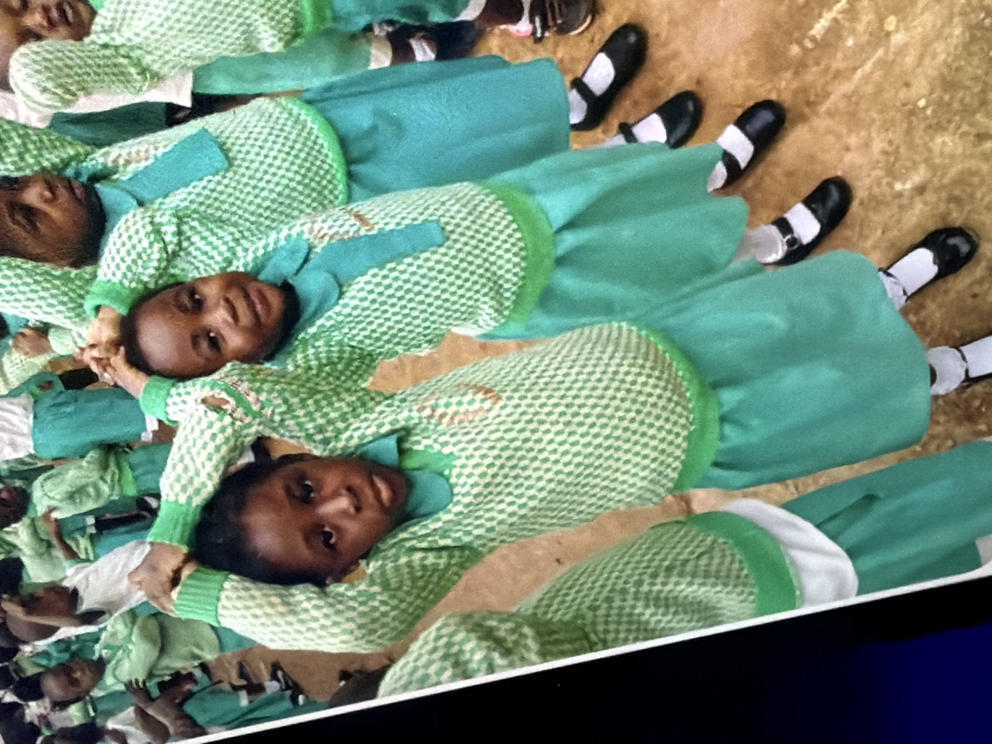 A group of young Black girls, dressed in matching green and white checkered sweaters and teal skirts with white socks and black shoes, stand in a line or cluster outdoors. The girls are of various ages, with some appearing to be toddlers and others pre-teens. They are all facing generally forward, with some looking at the camera. One girl in the foreground on the left has her hands raised above her head, and another girl in the center is also raising her hands. The background is blurred and appears to be dirt or sand, suggesting an outdoor setting such as a schoolyard or park. The lighting is natural and suggests daytime. The overall impression is one of a school or event.