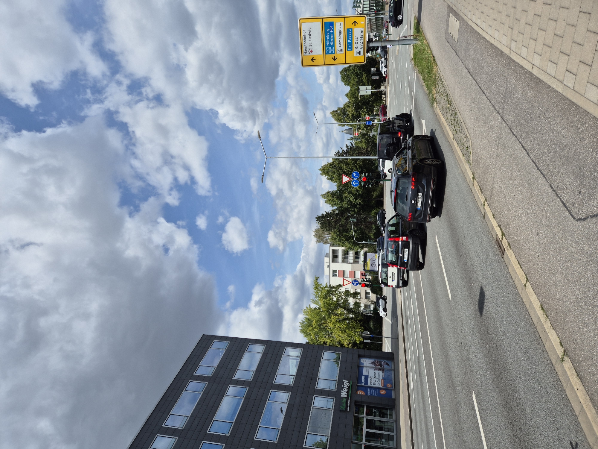 The image shows a street scene in Regensburg, Germany, with a modern, dark-colored office building on the left side of the frame. The building has numerous large, rectangular windows. In front of the building, a sign reads "Radio Weigl". Several cars are visible on the multi-lane road, including a black Volkswagen Passat wagon in the foreground. Traffic lights and road signs are present. One prominent yellow directional sign indicates "Hauptbahnhof St. Hedwig" with an upward arrow, "Nürnberg München-Hof" with a leftward arrow and a camping symbol, and "Passau R-Ost/GVZ/Hafen" with a rightward arrow. The sky is partly cloudy with a mix of gray and white clouds against a blue background. Trees and other buildings are visible in the background.