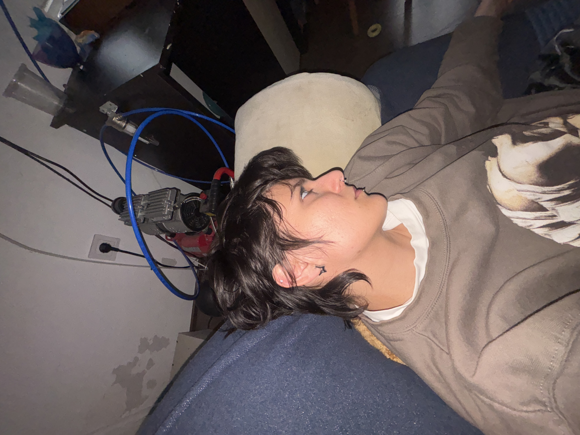 A person lies on a blue surface, appearing to be a couch or bed, while looking towards the upper right. The person wears a gray long-sleeve shirt over a white collar shirt. A tattoo is visible near the person's right ear. A beige cushion sits behind their head. To the left, a wall with some damp spots and an electrical outlet is visible, along with various wires and an air compressor. There is a desk above the compressor with various tools. The image provides no information that indicates the location in the city of Sofia, Bulgaria.