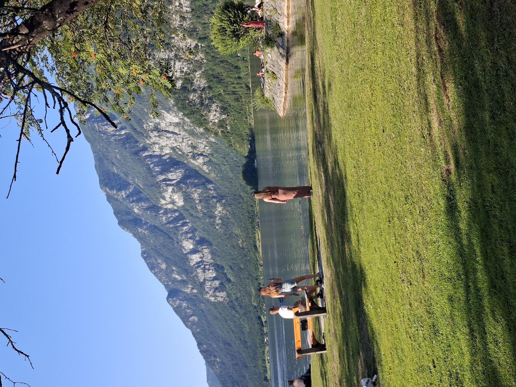 In Hallstatt, Austria, a tranquil scene unfolds by the serene lake, framed by majestic mountains. In the foreground, a vibrant green lawn slopes gently towards the water. A wooden bench, positioned on the grass, hosts two individuals: a man in a white t-shirt and shorts, and a woman in denim shorts and a t-shirt, both facing the lake. To the right of the bench, a woman in a flowing brown dress stands facing away from the camera, gazing towards the water. Further along the shore, a rocky outcrop features a group of people, some seated, enjoying the picturesque setting. The lake's calm surface reflects the surrounding lush greenery and the imposing, rugged mountain range that dominates the horizon. The sky above is a clear, brilliant blue, with the branches of a tree, adorned with orange berries, extending into the frame from the upper right. The overall atmosphere is one of peaceful contemplation and immersion in natural beauty.