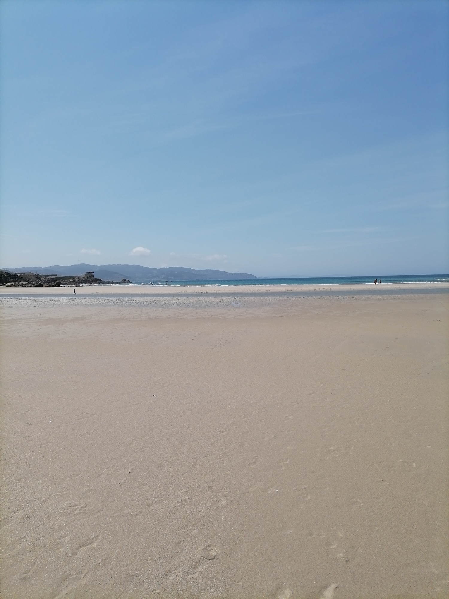 The image depicts a wide, sandy beach under a clear blue sky with a few faint clouds. The foreground is dominated by the texture of the damp sand, showing some footprints and subtle ripples. In the midground, a shallow body of water reflects the sky, suggesting it is low tide or a tidal pool. Beyond this, the ocean stretches to the horizon, with small waves breaking near the shore. In the distance, a low, hilly coastline is visible, dotted with some vegetation and rocky outcrops. There are a few scattered people visible on the beach and in the water. On the left side of the midground, a lone figure walks along the sand. Further to the right, near the water's edge, two figures are visible, one appearing to be carrying a surfboard. The overall impression is one of a peaceful, expansive coastal environment, likely during daylight hours. Given the context of Arteixo, Spain, this scene is likely representative of one of its coastal areas such as Praia de Area Grande or Praia de Valcobo.
