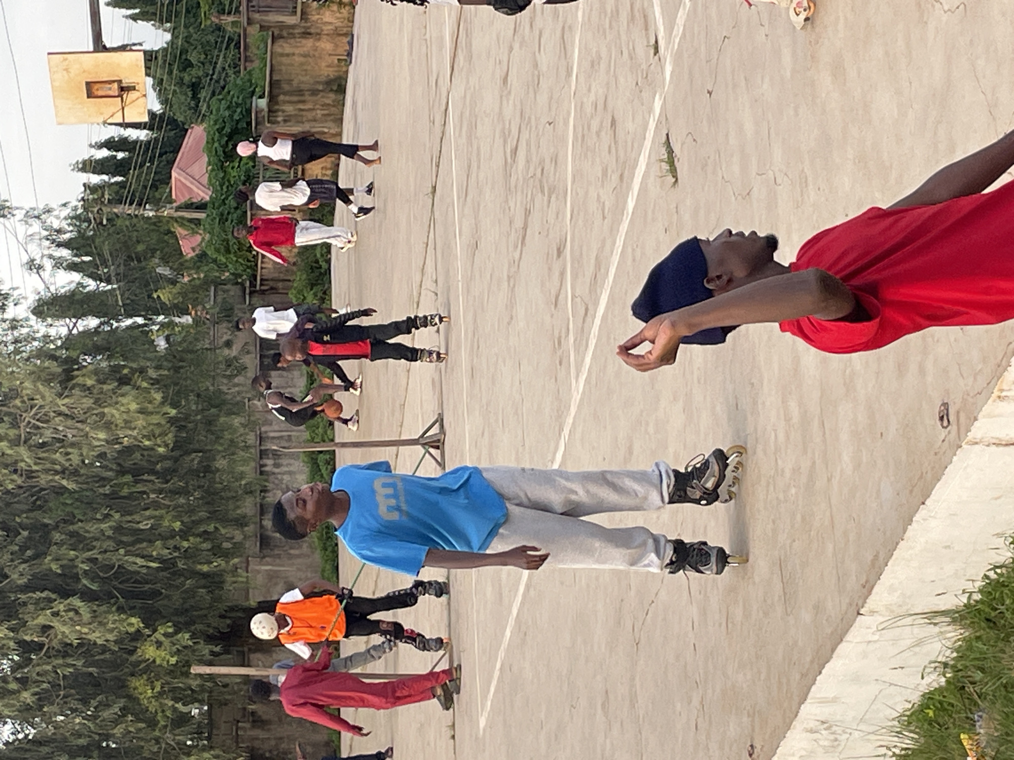In Jos, Nigeria, on a sun-drenched outdoor basketball court, several young people engage in rollerblading and basketball. In the foreground, a young man on rollerblades wearing a blue t-shirt and gray sweatpants stands facing away from the camera. To his left, another young man in a red tracksuit on rollerblades is visible. Further back, a group of individuals are playing basketball, one of whom is wearing an orange vest and white helmet. The court, marked with white lines, is surrounded by a wall and lush greenery, with trees and power lines visible in the background.
