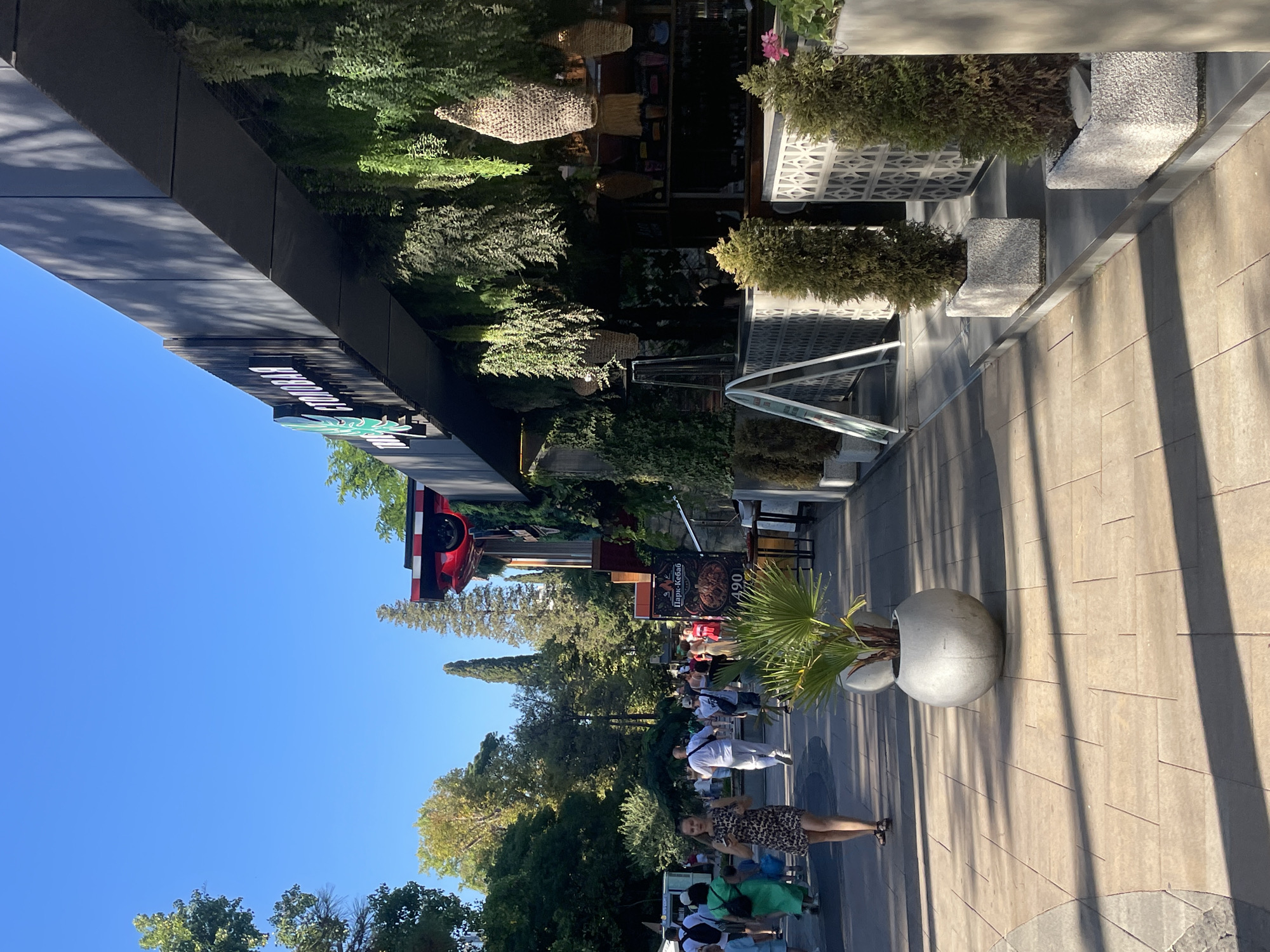 This is a medium shot, taken from a slightly low angle, on a sunny day in the city of Sochi, Russia. The scene captures a lively pedestrian walkway in front of a modern building adorned with lush greenery and hanging plants. The walkway is paved with light-colored tiles, with distinct shadows cast by unseen trees, suggesting it is late afternoon.
In the foreground, a woman with dark hair, wearing a patterned dress and sandals, is walking away from the camera, holding a phone in her right hand. To her left, a child in a blue outfit is walking. Further down the walkway, a crowd of people, dressed in casual summer clothing, are strolling in both directions. Some are carrying bags, and others are engaged in conversation.
The building on the right features a dark facade with a prominent sign that reads "СТОЛОВАЯ" (Stolovaya, meaning canteen or cafeteria) in white, illuminated letters. This building is heavily decorated with hanging plants, creating a natural, jungle-like effect. Several woven baskets are suspended from the building's overhang.
To the right of the walkway, there are planters containing cypress trees in modern, grey containers. A sign with the text "Парк-Кебаб" (Park-Kebab) and a price of "490" is visible on the building. In the background, tall green trees and cypress trees create a verdant backdrop, contrasting with the clear blue sky. The overall atmosphere is one of a bustling, outdoor public space on a warm day.