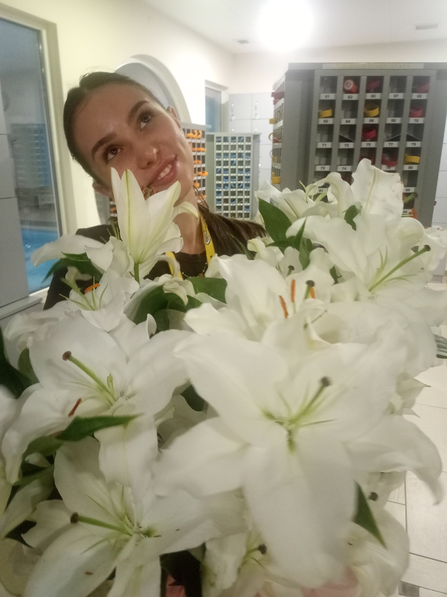 A woman is holding a large bouquet of white lilies in front of her. She is smiling and looking up. The background appears to be a locker room or a similar facility with rows of lockers and cabinets. The lighting is bright, suggesting an indoor setting.