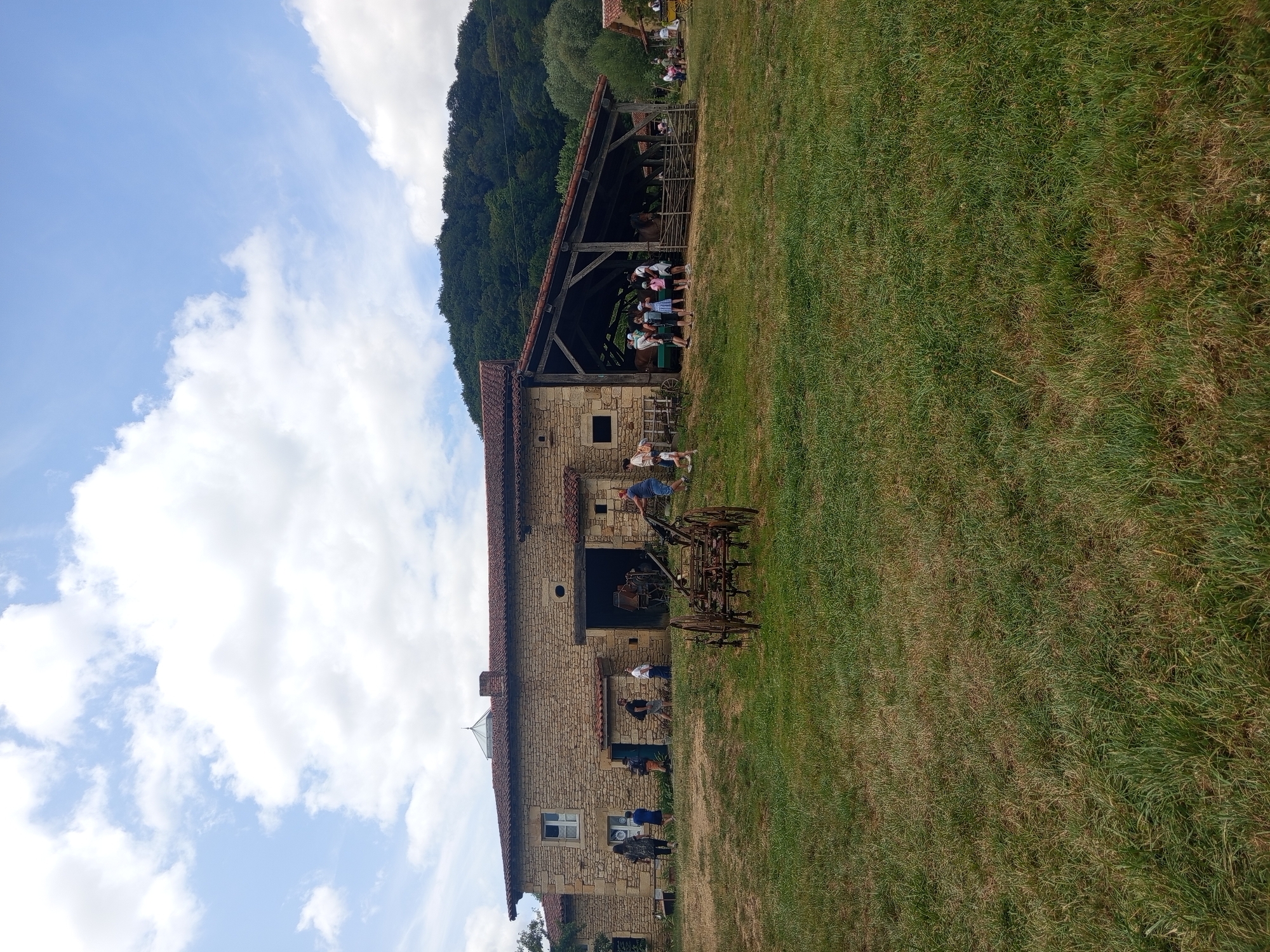 The image captures an outdoor scene at a historical reenactment or heritage site in Azannes-et-Soumazannes, France, on a partly cloudy day. A stone building with a red-tiled roof dominates the background, featuring an open-sided covered area where several people, including children, are gathered. Two horses are visible within this covered area, behind a wooden fence. In the foreground, a group of individuals, seemingly visitors, are observing the site and interacting. Various antique farming implements are displayed, including a weathered wooden and metal plow in the middle ground and other machinery near the building. The environment consists of a grassy slope leading up to the structures, with a treed hillside forming the backdrop under a sky with scattered white clouds.