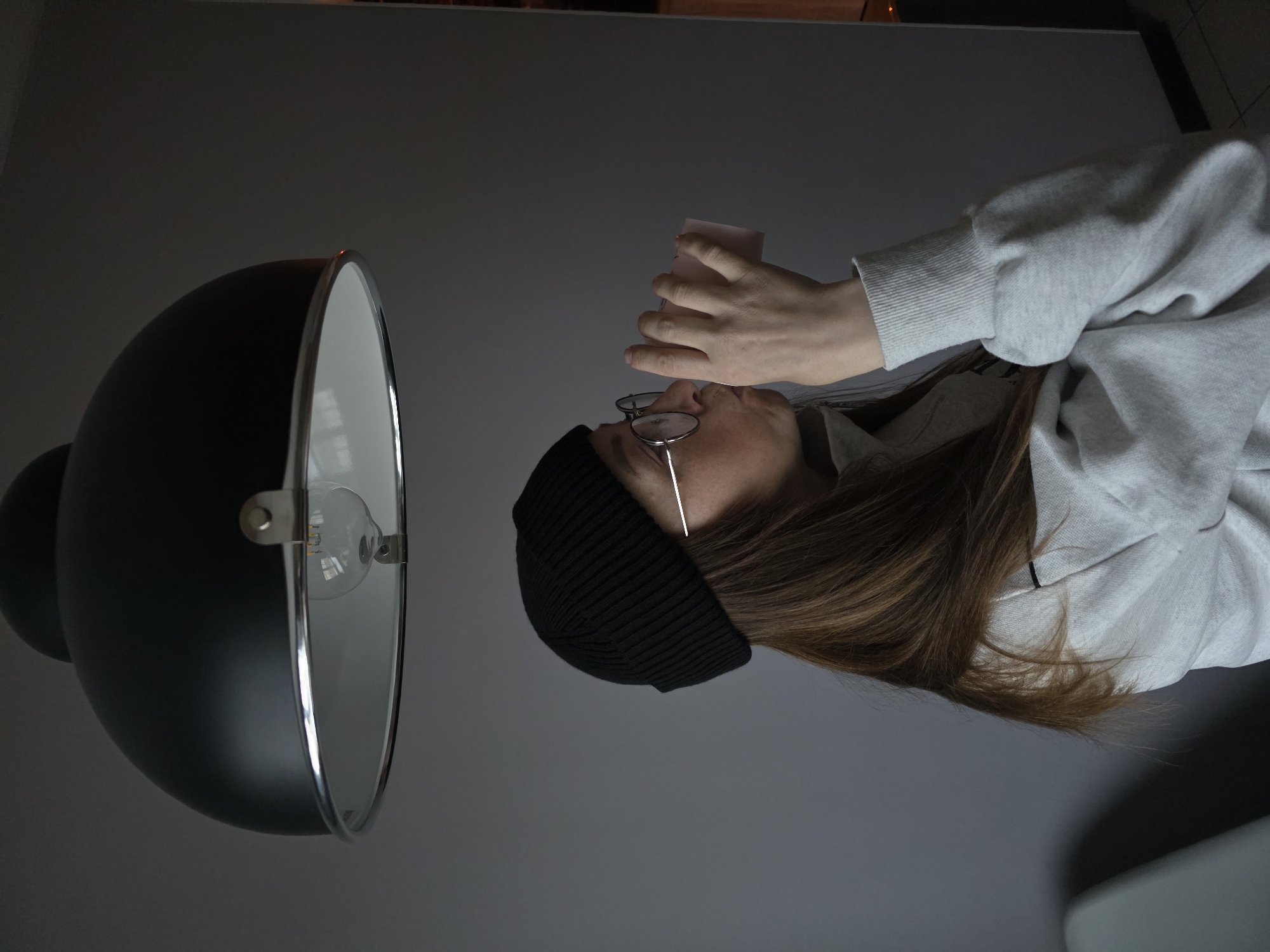A person wearing a black beanie and glasses is holding a small object near their face, positioned next to a large black lamp with a white interior. The person is wearing a light grey sweatshirt. The background is a plain grey wall. The image was likely taken indoors. The city of Chaikovsky, Russia, cannot be verified as the location based solely on the provided image.