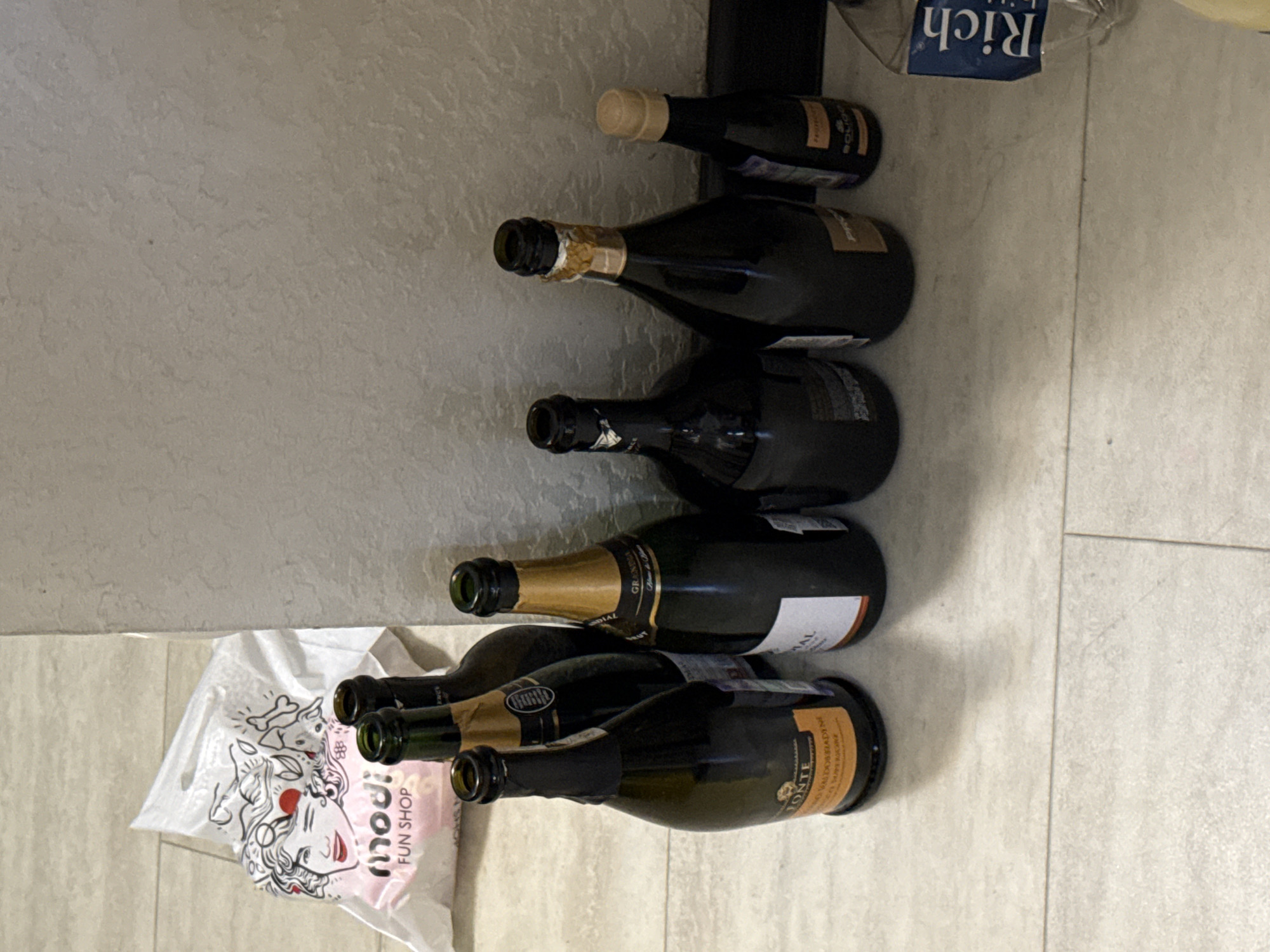 The image shows a wine rack with six bottles of wine, placed against a light-colored wall and floor. There are a few objects near the wine rack, including a plastic bag with a logo that says "Modi fun shop" on the bottom left of the image and a "Rich" box on the top right of the image. The location in the city of Sochi, Russia, cannot be determined from the image.