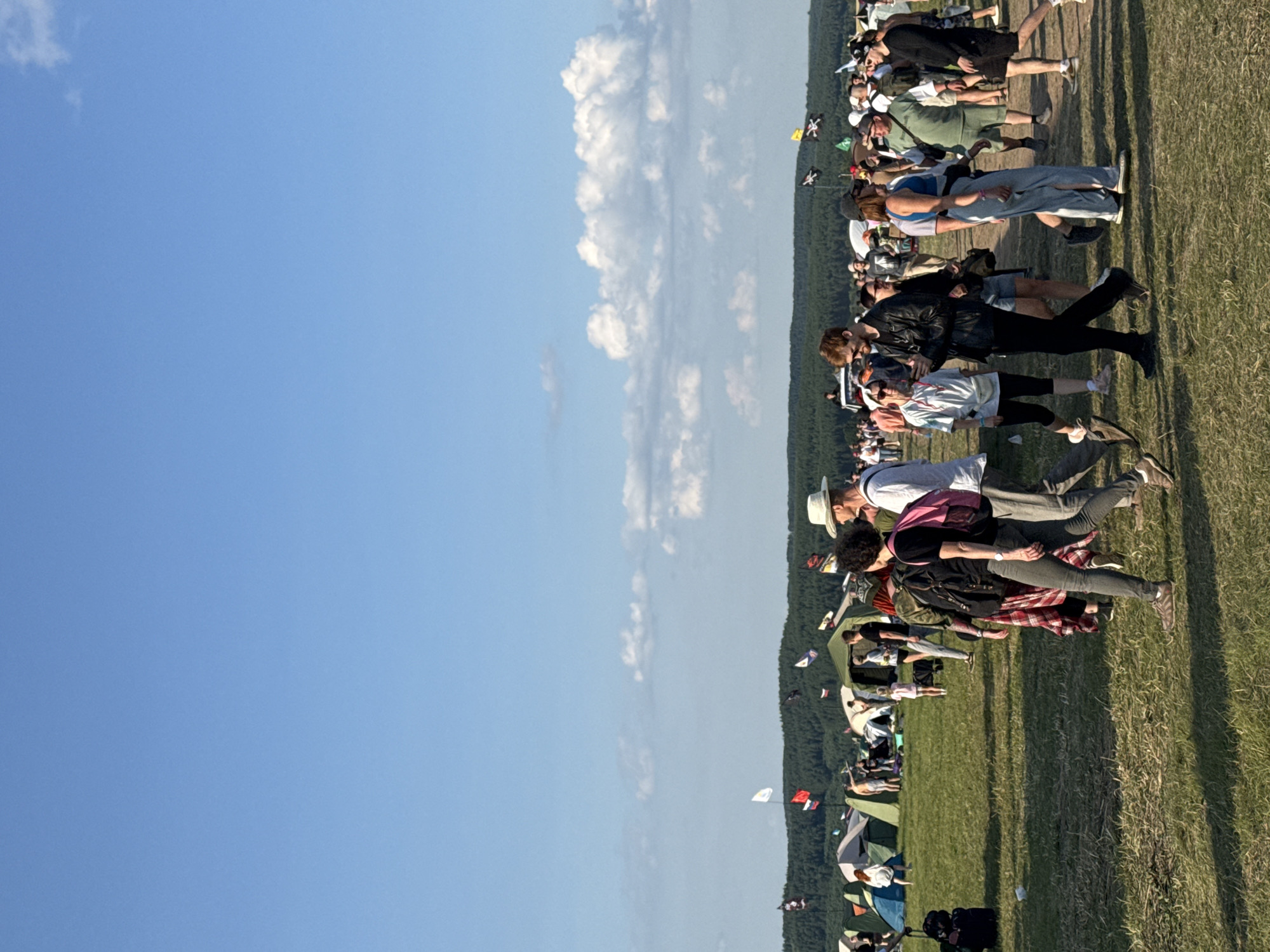 The image depicts an outdoor scene at what appears to be a festival or campsite under a clear blue sky with scattered clouds. Numerous tents are set up on a grassy field, and a line of trees forms a backdrop on a distant hill. Several people are visible in the foreground and midground, walking and interacting. Some are dressed casually in t-shirts and shorts, while others are wearing more distinctive attire like a white hat and a leather jacket. Various flags, some with pirate motifs, are planted in the field. The overall atmosphere suggests a daytime event.