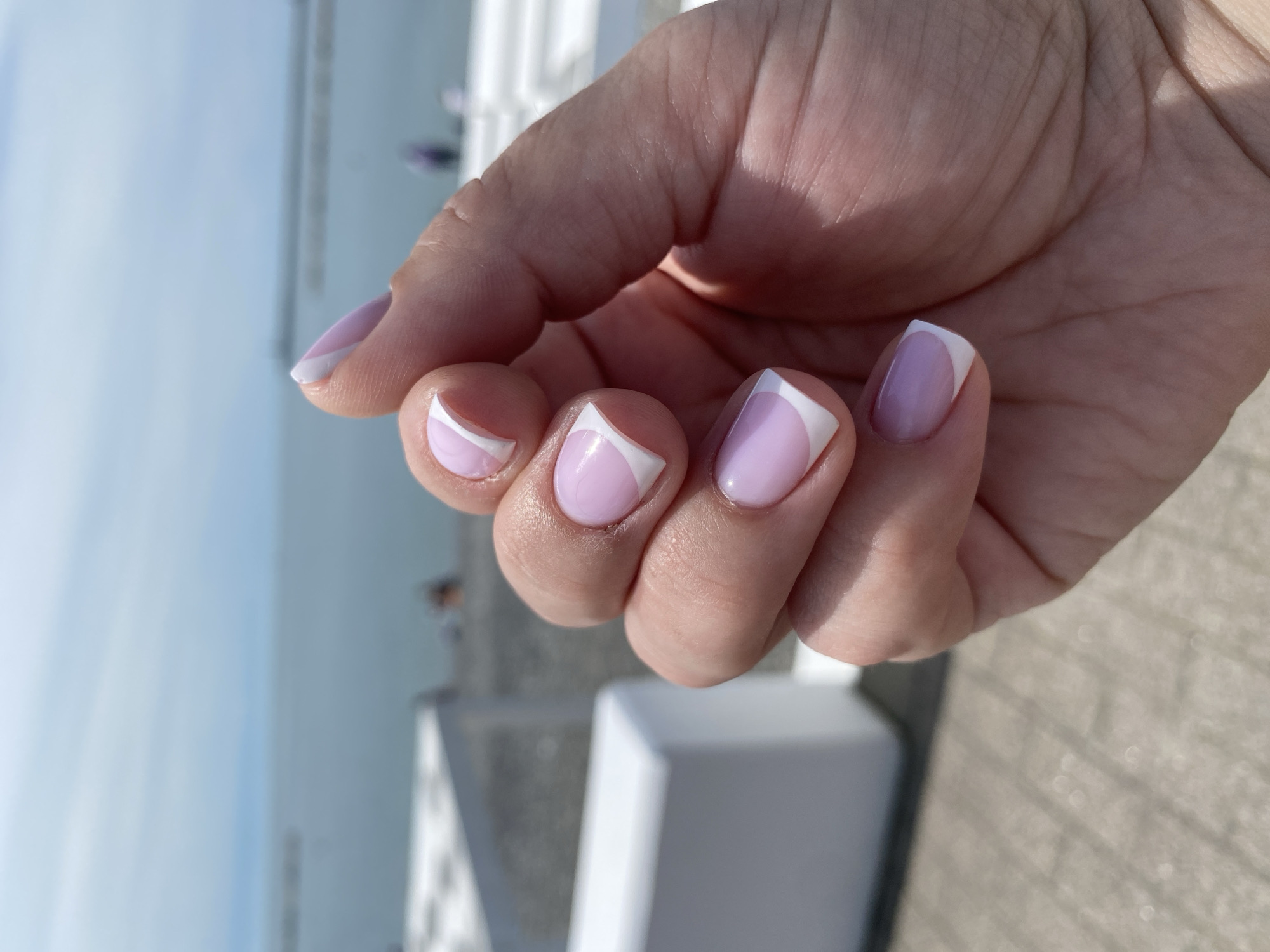 A close-up shot shows a person's hand with a fresh manicure. The nails are painted a pale purple color, with a geometric white French tip design. The hand is slightly curled, with the fingers bent towards the palm. The background is blurred, but suggests an outdoor setting with a white structure to the left and a grey concrete surface below. The location is not discernible as Sochi, Russia.