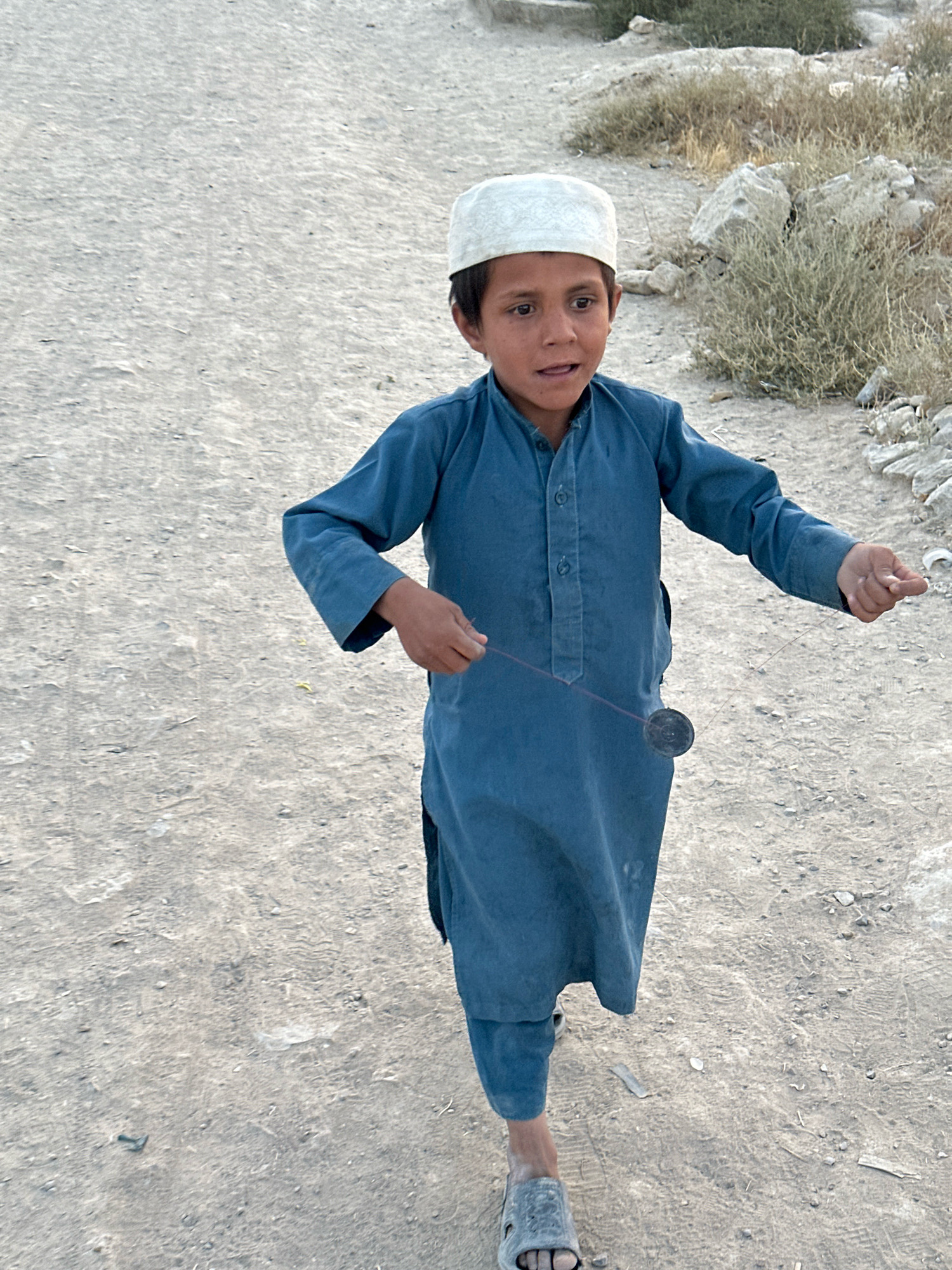 A young boy in Kuchlak, Pakistan, is captured mid-motion, engaged in playing with a yo-yo. He is dressed in a traditional blue shalwar kameez and a white kufi cap. The boy's facial expression suggests concentration and perhaps excitement as he plays. The ground beneath him is unpaved, dusty, and scattered with small stones and dry vegetation, indicative of an outdoor environment in a dry or arid region. The background shows more of this natural, sparsely vegetated terrain with larger rocks. The lighting suggests it is daytime, possibly late afternoon given the soft shadows.