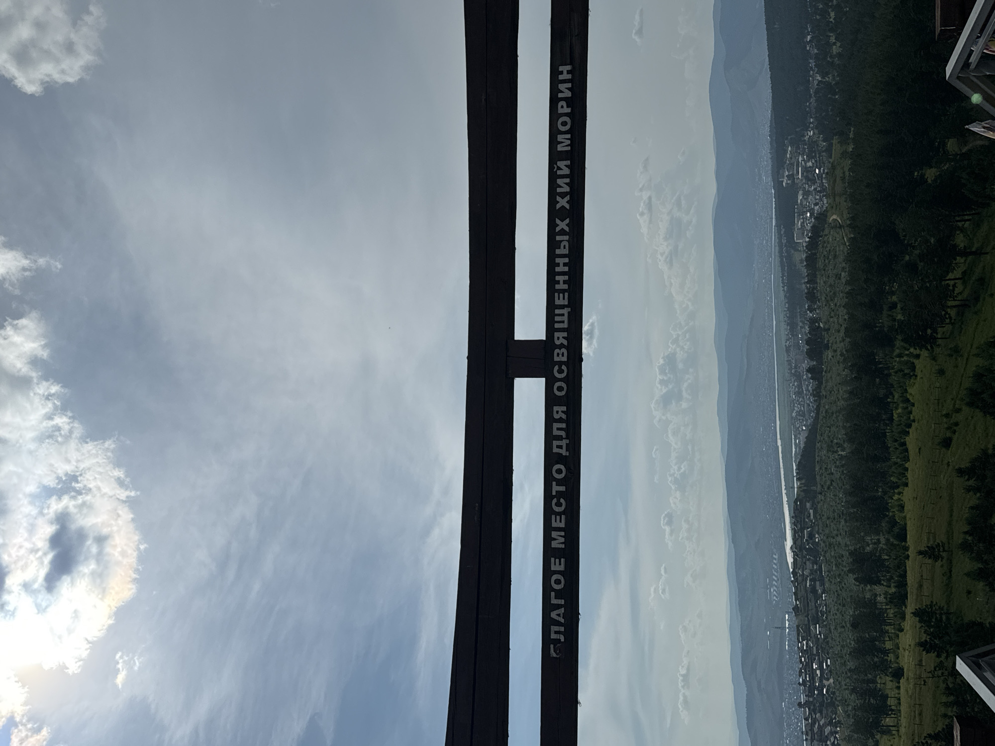 The image shows a panoramic view from an elevated vantage point overlooking the city of Ulan-Ude, Russia. In the foreground, a dark wooden railing or structure is visible, with text inscribed on it. The text appears to be in Cyrillic script and reads "Благое место для освященных хий морин". This translates to "A good place for consecrated sky horses" or "A sacred place for consecrated sky horses".
Beyond the foreground structure, the landscape unfolds to reveal a sprawling city nestled within a valley. Numerous buildings, ranging from residential areas with smaller structures to larger, more modern buildings, are visible. A river winds through the city, reflecting the sky. Rolling hills and mountains form a distant backdrop, fading into a hazy blue due to atmospheric perspective. The sky is partially cloudy with patches of sunlight breaking through, creating a dynamic interplay of light and shadow on the landscape below. The overall impression is one of a scenic overlook offering a broad perspective of the urban and natural environment of Ulan-Ude. There are no people or animals visible in the frame.