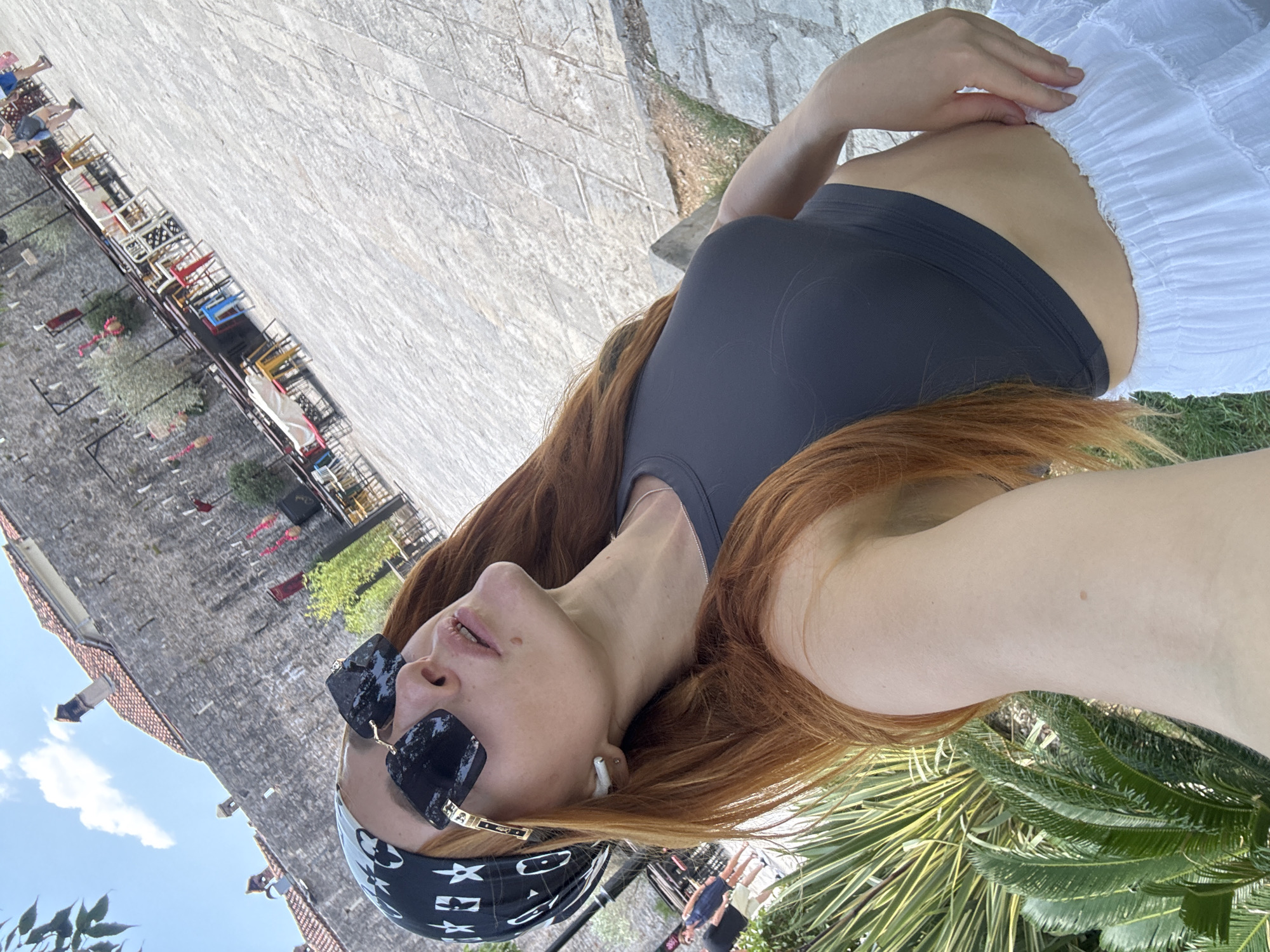 The image captures a selfie taken by a woman with long, auburn hair. She is wearing a black bandana with a white pattern, large square sunglasses with dark lenses, and a dark gray crop top. She also has a white wireless earbud in her left ear and is wearing a white ruffled skirt. In the background, a stone wall of an old building or fortress is visible, with red-tiled roofs and a clear blue sky with some clouds. Several tables and chairs, some colorful, are set up in an outdoor area in front of the wall. A few people can be seen in the distance, walking in the paved outdoor area. The woman is standing on a paved surface with some greenery and foliage in the foreground. The scene appears to be in a historical or tourist area of Budva, Montenegro, possibly near the Old Town walls, given the architectural style and the presence of outdoor seating.