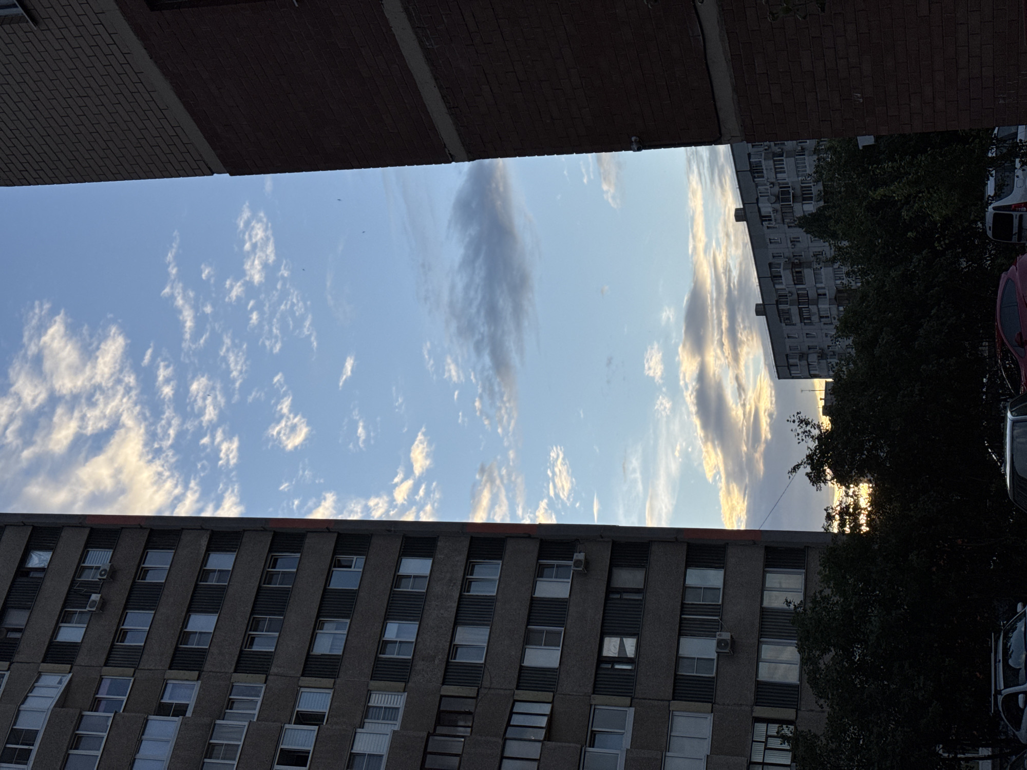 An outdoor, eye-level, full shot captures a sliver of sky between two buildings. On the left, a grey, multi-story apartment building with many windows is visible. Air conditioning units are mounted on some of the windows. On the right, a portion of a red brick building is shown, with a lighter band of concrete separating the upper and lower sections. The sky is blue with scattered white clouds and some darker, larger clouds. In the distance, another apartment building with a similar architectural style to the one on the left can be seen. The foreground is dominated by dark green trees, obscuring the lower parts of the buildings. At the very bottom, the hoods of parked cars are visible. The lighting suggests it is either early morning or late afternoon, with the sun casting a warm glow on some of the clouds.
